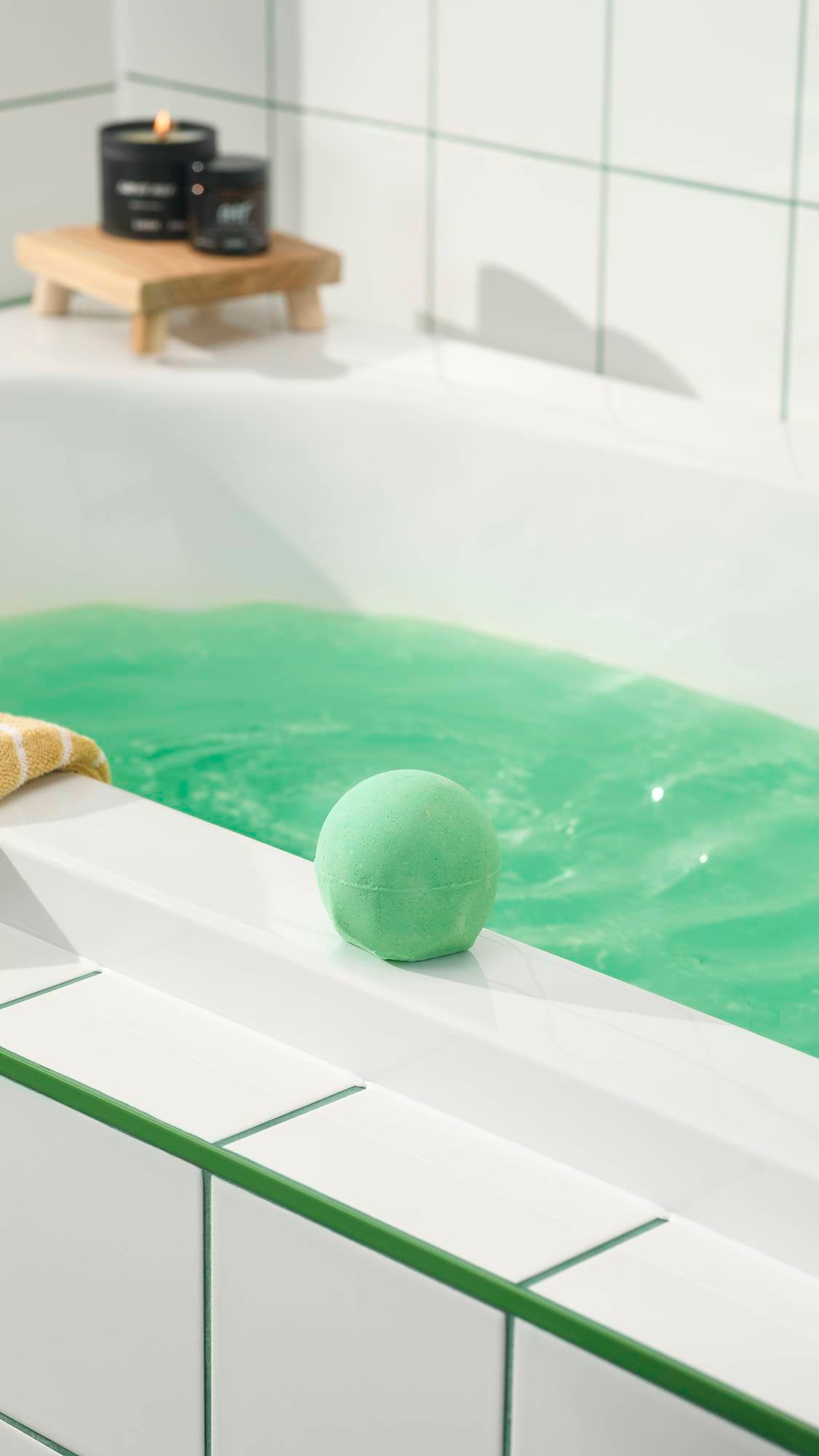 The green Avobath Bath bomb sits on the white edge of a bathtub filled with shimmering green water. In the background, lit candles rest on a small wooden stand against a white tiled wall.