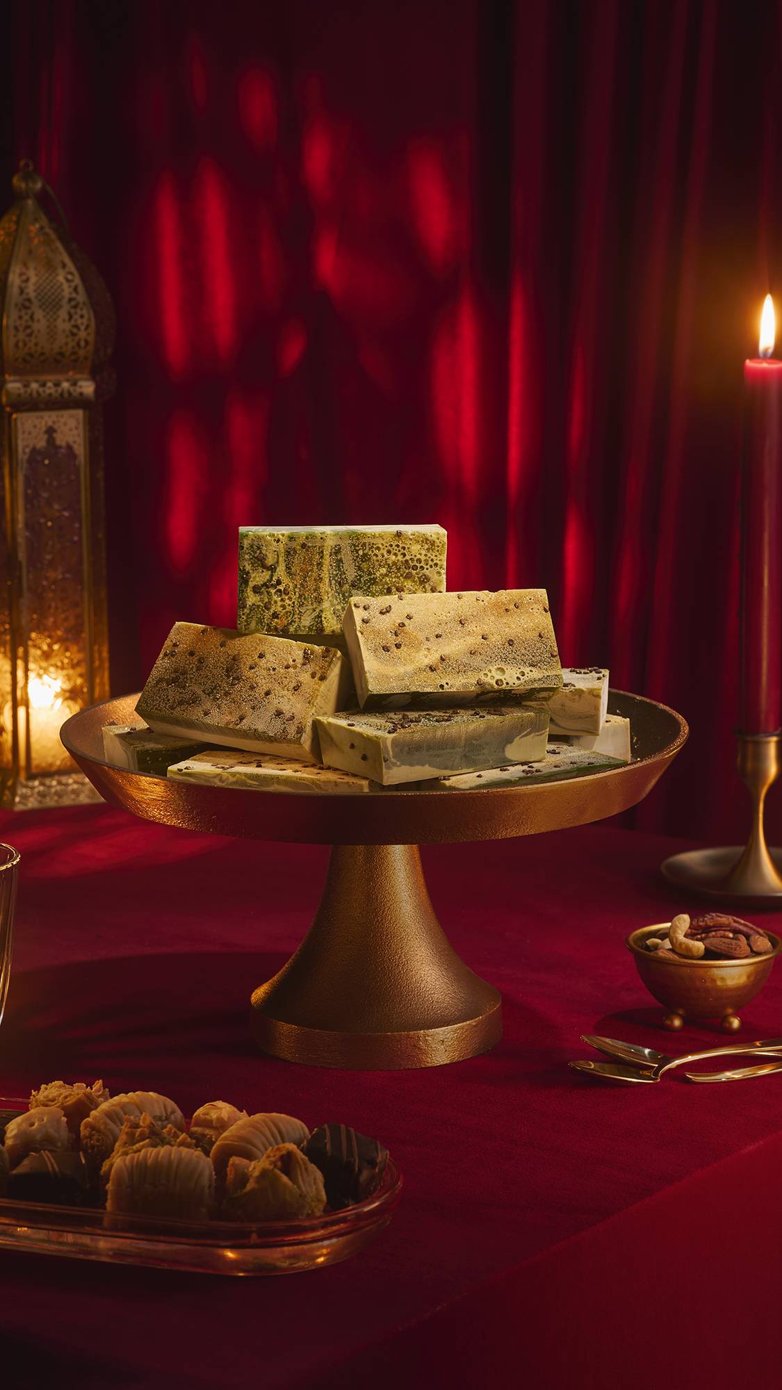 A stack of marbled green "Baklava" soap bars on a brass cake stand. The festive scene includes a red velvet tablecloth, a lit candle, an ornate gold lantern, and plates of baklava and nuts against a backdrop of draped red fabric.