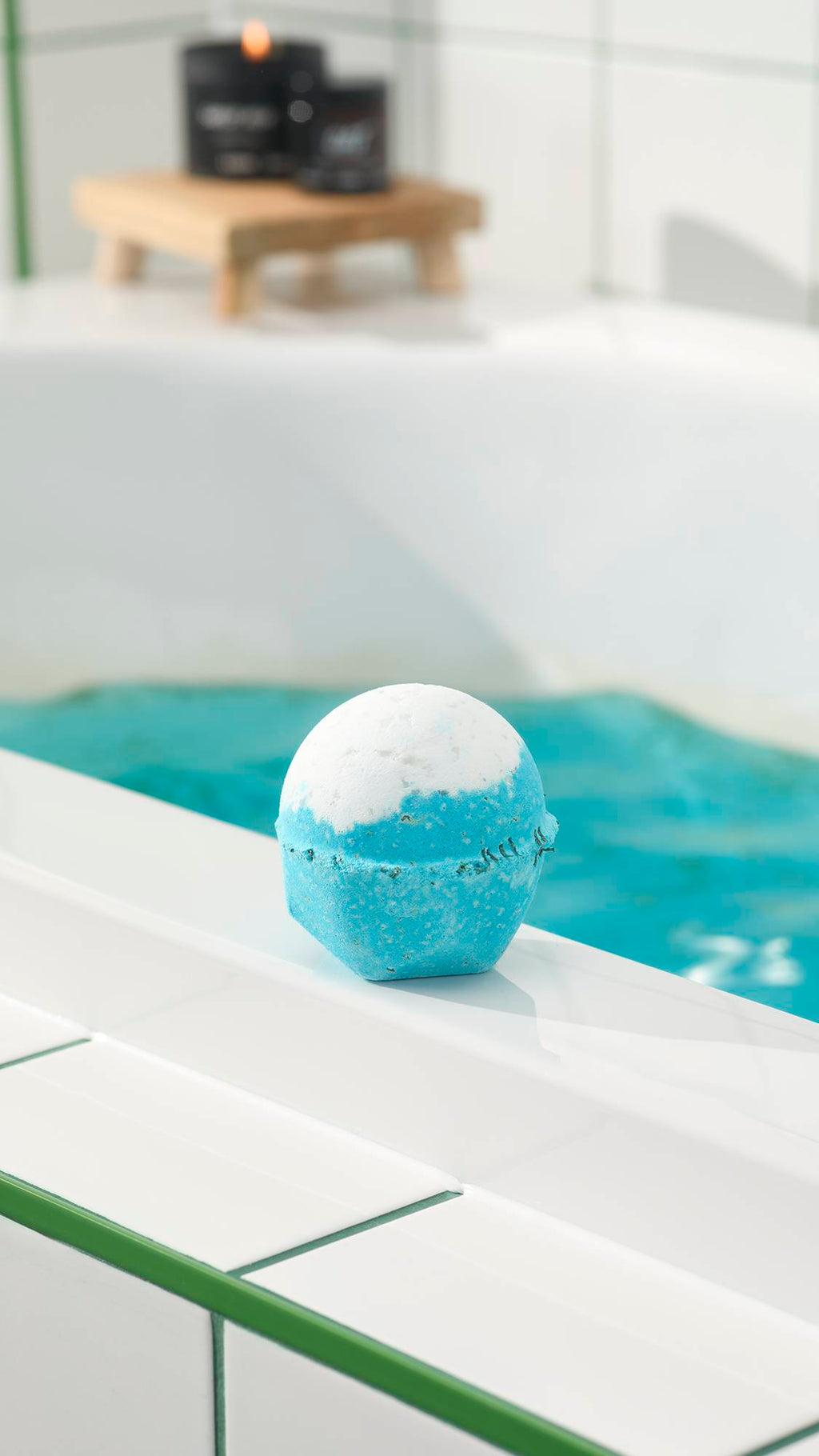 The blue and white Big Blue bath bomb sits on the white edge of a bathtub filled with bright blue water. In the background, a lit candle rests on a wooden stand against white tiles.
