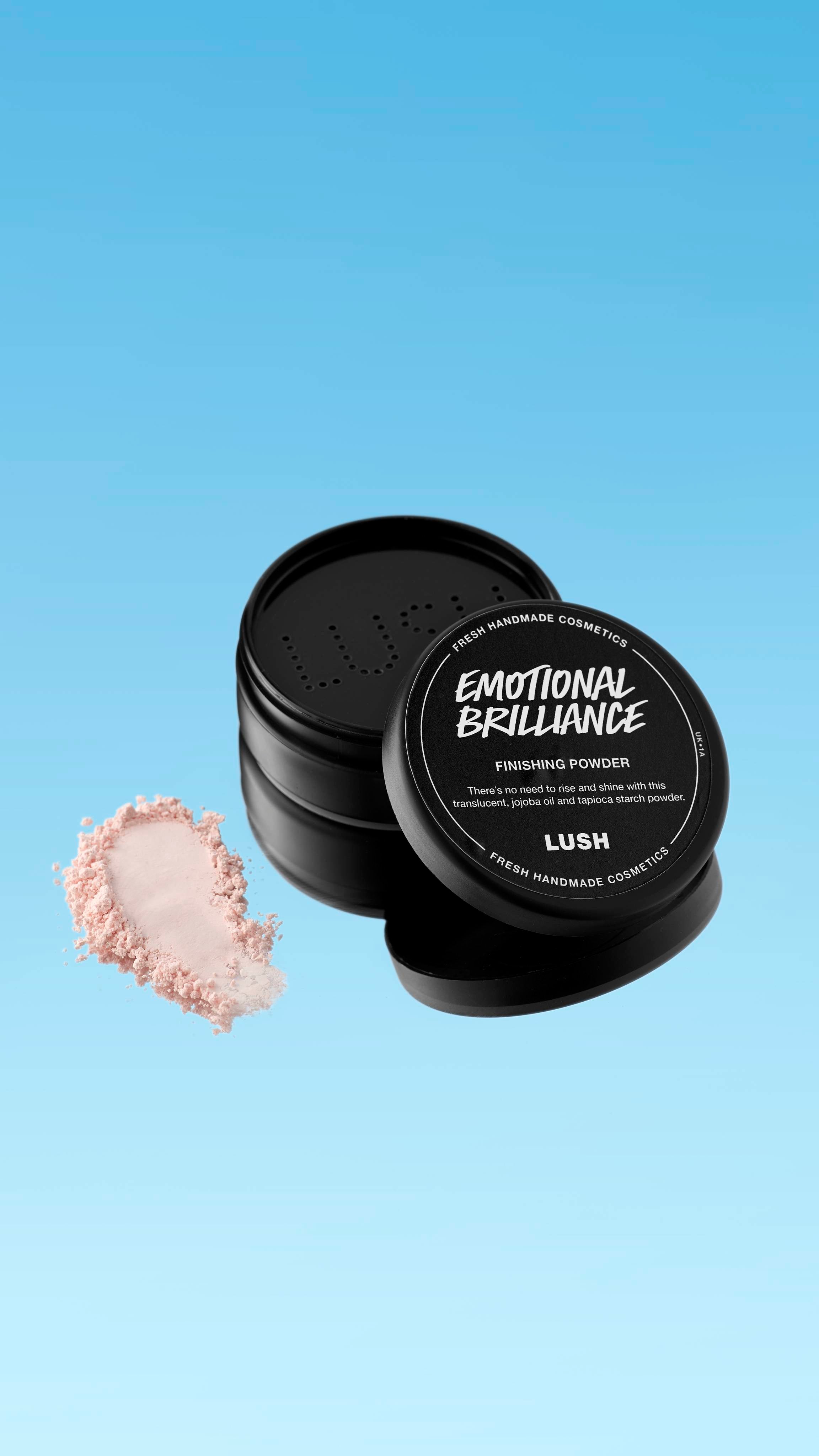 A partially open black pot of Emotional Brilliance ~ Finishing Powder is shown floating against a blue gradient background. A small pile of the pale pink powder is visible next to the pot's base. The black lid has white text identifying the product.