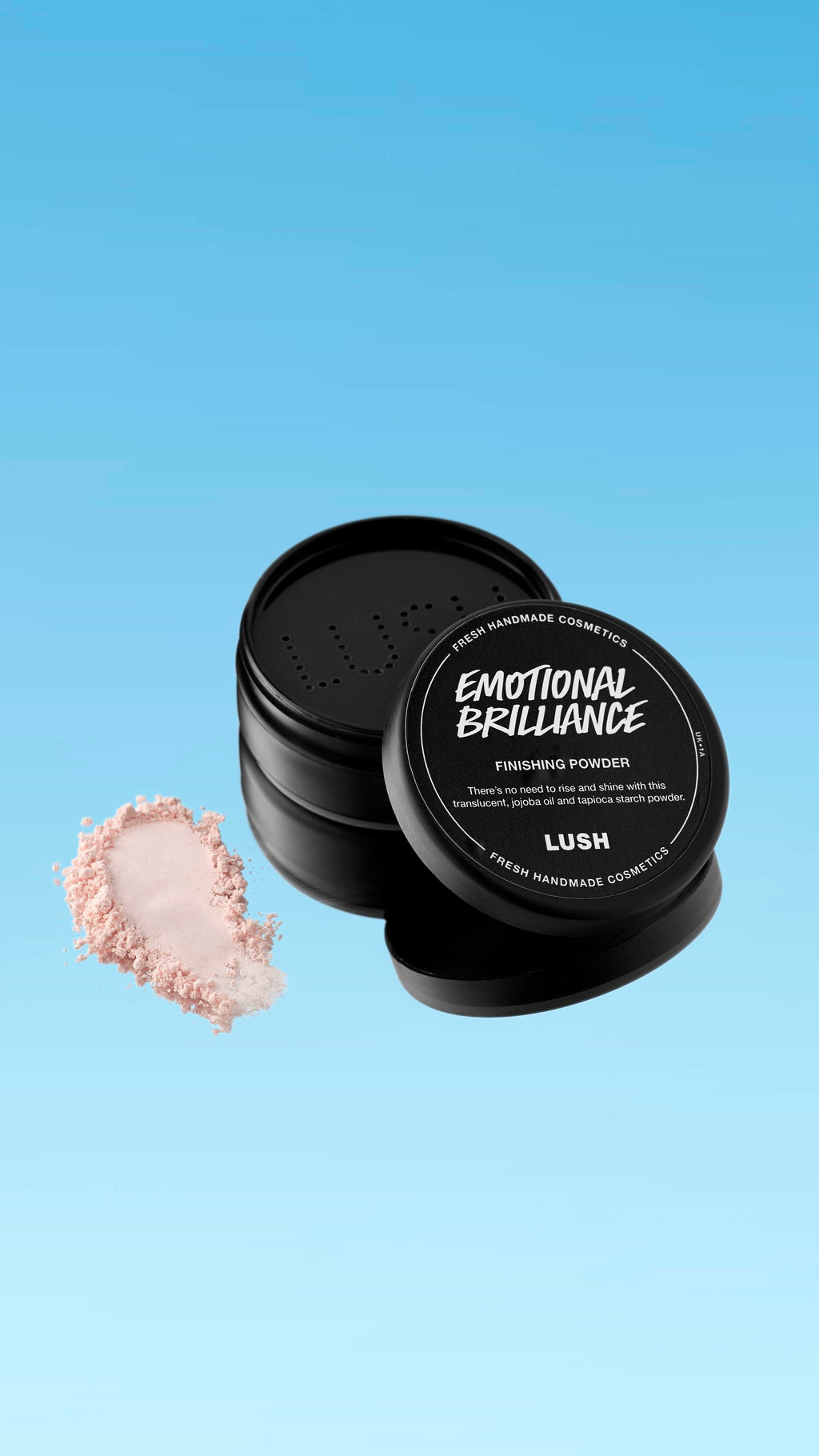 A partially open black pot of Emotional Brilliance ~ Finishing Powder is shown floating against a blue gradient background. A small pile of the pale pink powder is visible next to the pot's base. The black lid has white text identifying the product.
