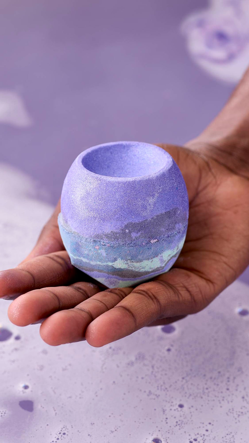 The image shows a hand holding a round, purple "Goddess" bath bomb with a grey and mint green marbled base over a tub of lilac-coloured bathwater. The bath bomb has a bowl-shaped indentation on top.