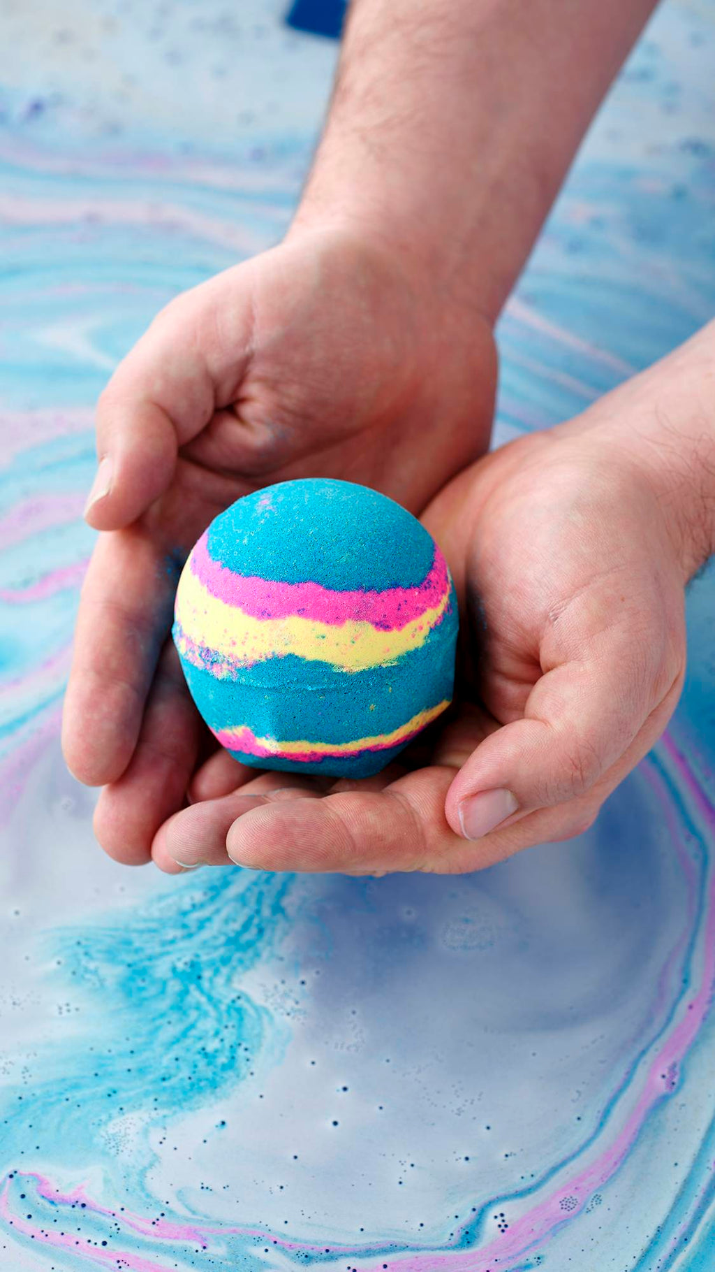 A pair of hands holds a spherical Intergalactic bath bomb over blue and pink swirling water. The bomb is primarily bright blue with vibrant horizontal stripes of neon pink and yellow.