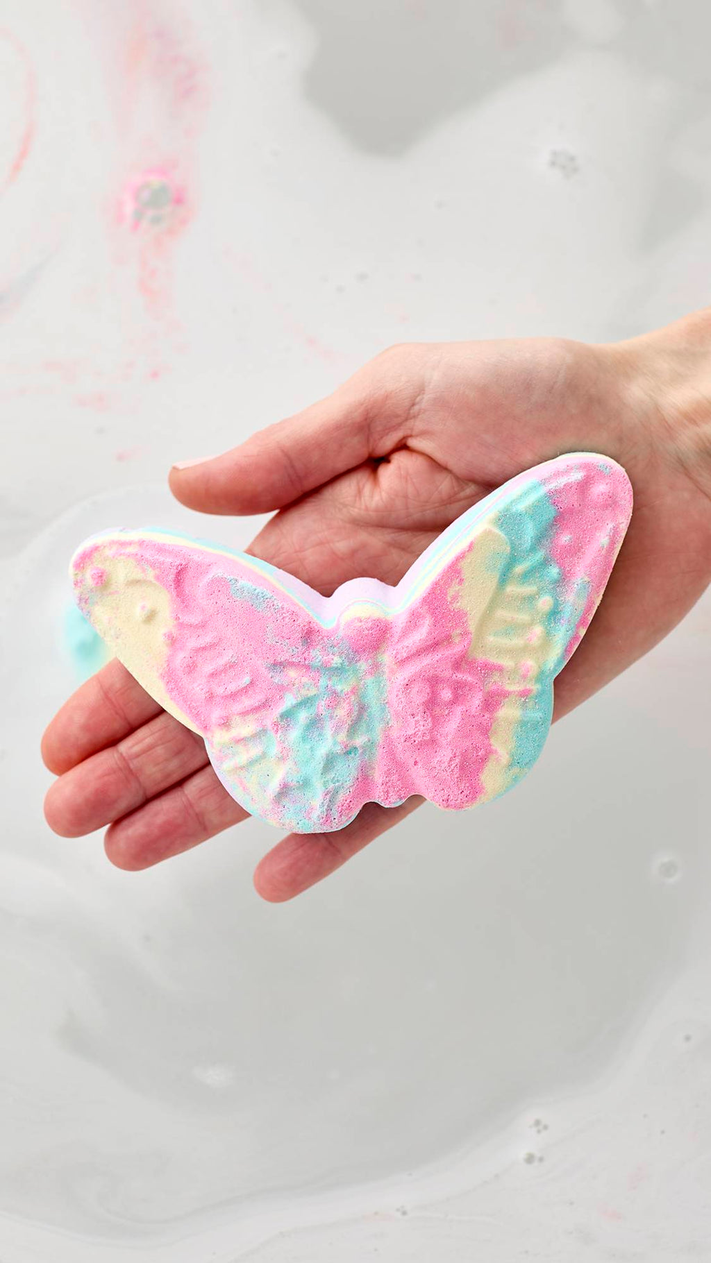 A hand holds a butterfly-shaped bath bomb with marbled swirls of pink, yellow, and turquoise over a bath of milky white water. Faint streaks of pink color are visible in the background as the bomb begins to dissolve.