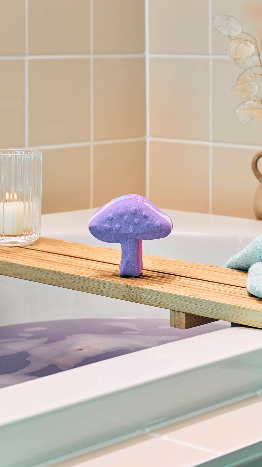 A purple "Magik Mushroom" bath bomb sits upright on a wooden bath tray over a tub of lilac-coloured water. In the background, beige tiles and a lit candle in a glass holder create a serene atmosphere.