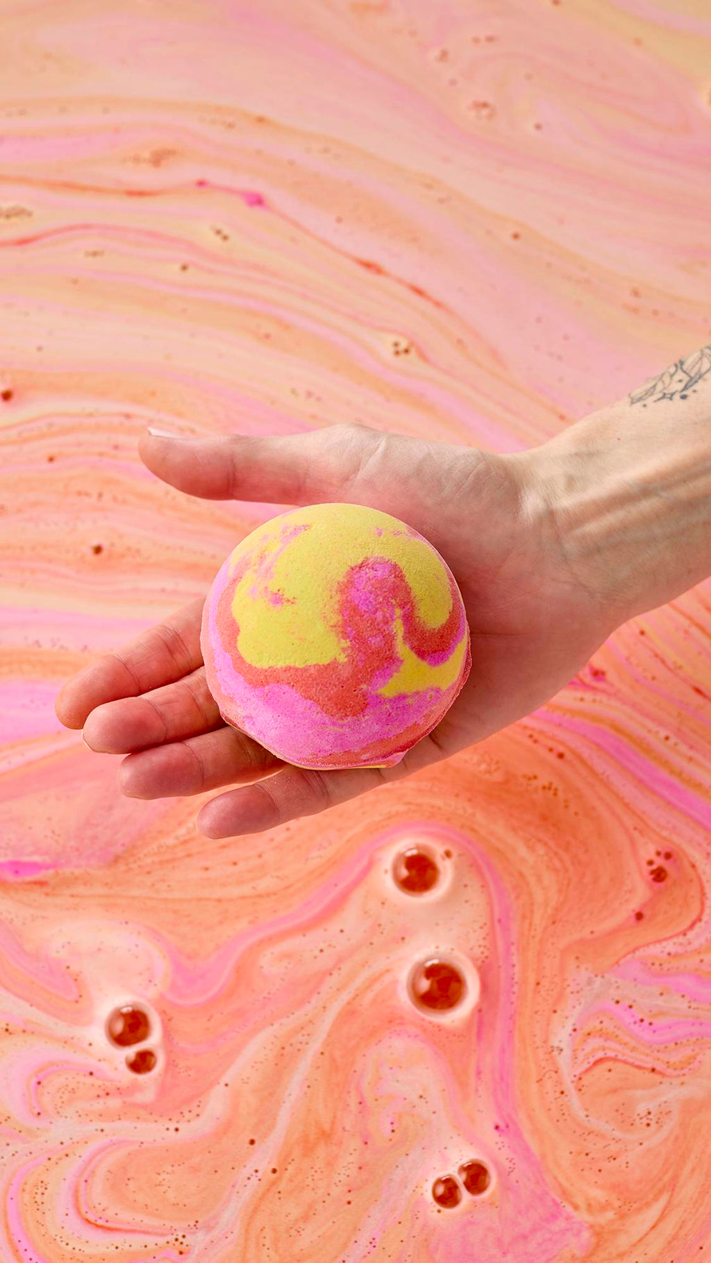 A hand holds a vibrant yellow, pink, and orange marbled bath bomb over a bath of swirling peach-colored water. Pink and orange foam patterns create a marble-like texture across the water's surface as the bomb begins to dissolve.