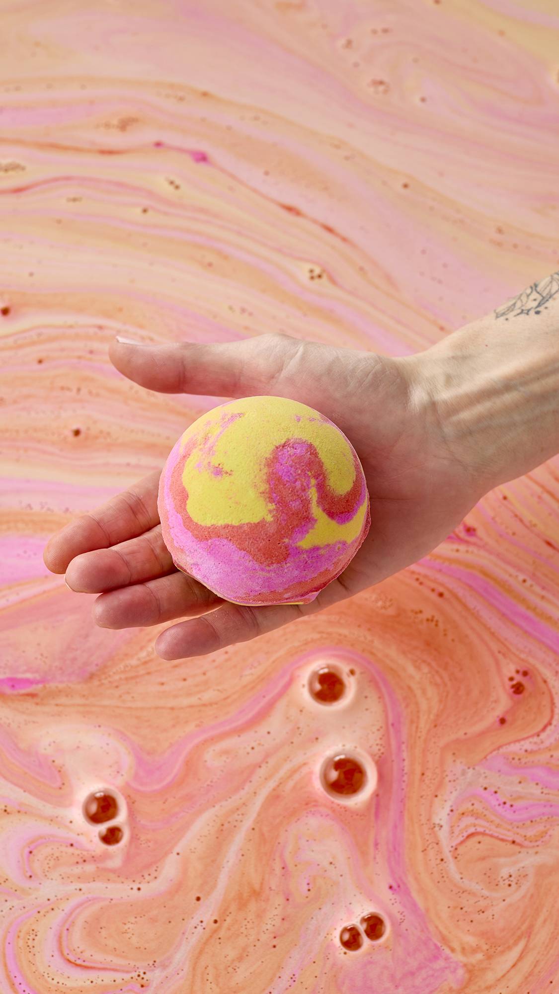 A hand holds a vibrant yellow, pink, and orange marbled bath bomb over a bath of swirling peach-colored water. Pink and orange foam patterns create a marble-like texture across the water's surface as the bomb begins to dissolve.
