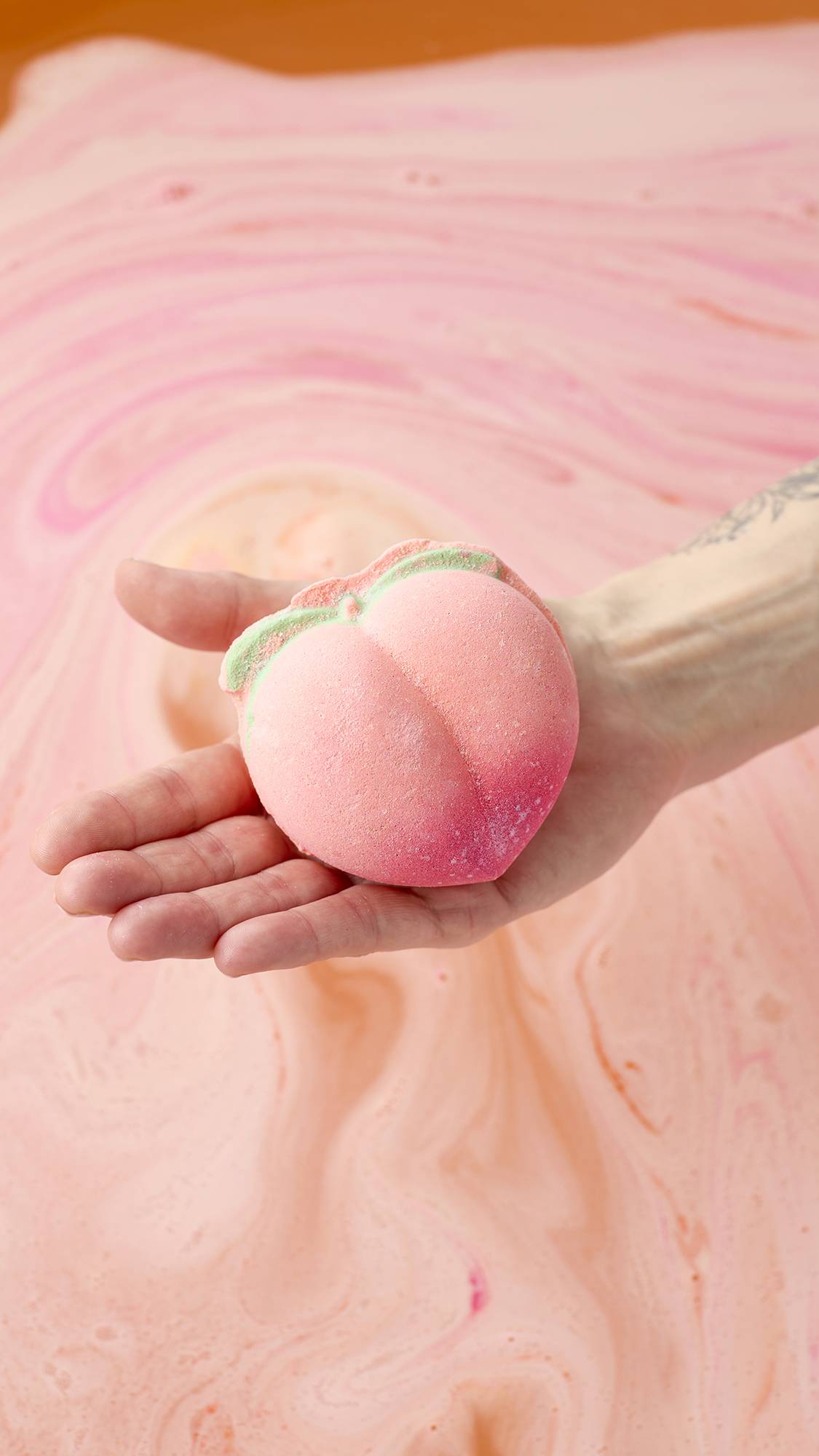 A hand holds a peach-shaped bath bomb over a tub of swirling peach and pink bathwater. The bath bomb has a soft pink gradient and small green leaves at the top. The water surface is covered in delicate foam and vibrant streaks of colour.