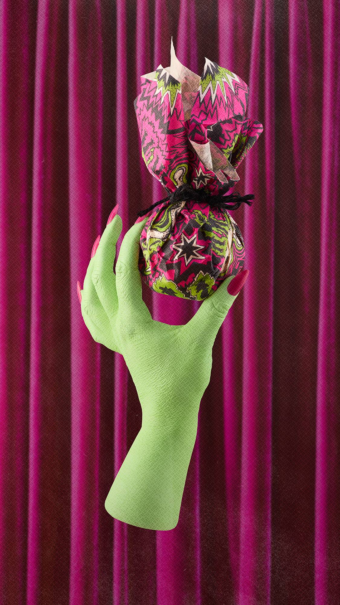 A lime green hand with long pink fingernails holds up a small gift wrapped in a vibrant magenta, black, and green patterned "Screech" Lokta paper, tied with a black string. The hand is set against a background of draped magenta velvet curtains.