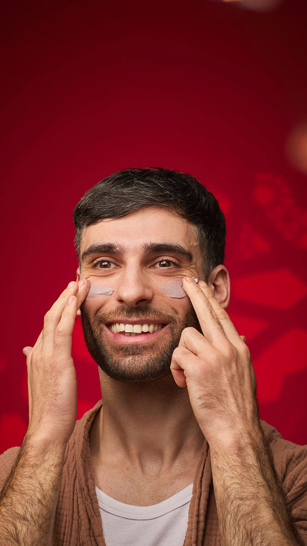 A person smiles while applying dabs of the mauve Sipak face mask to their cheekbones. They wear a brown textured robe against a deep red background with soft golden bokeh lights.