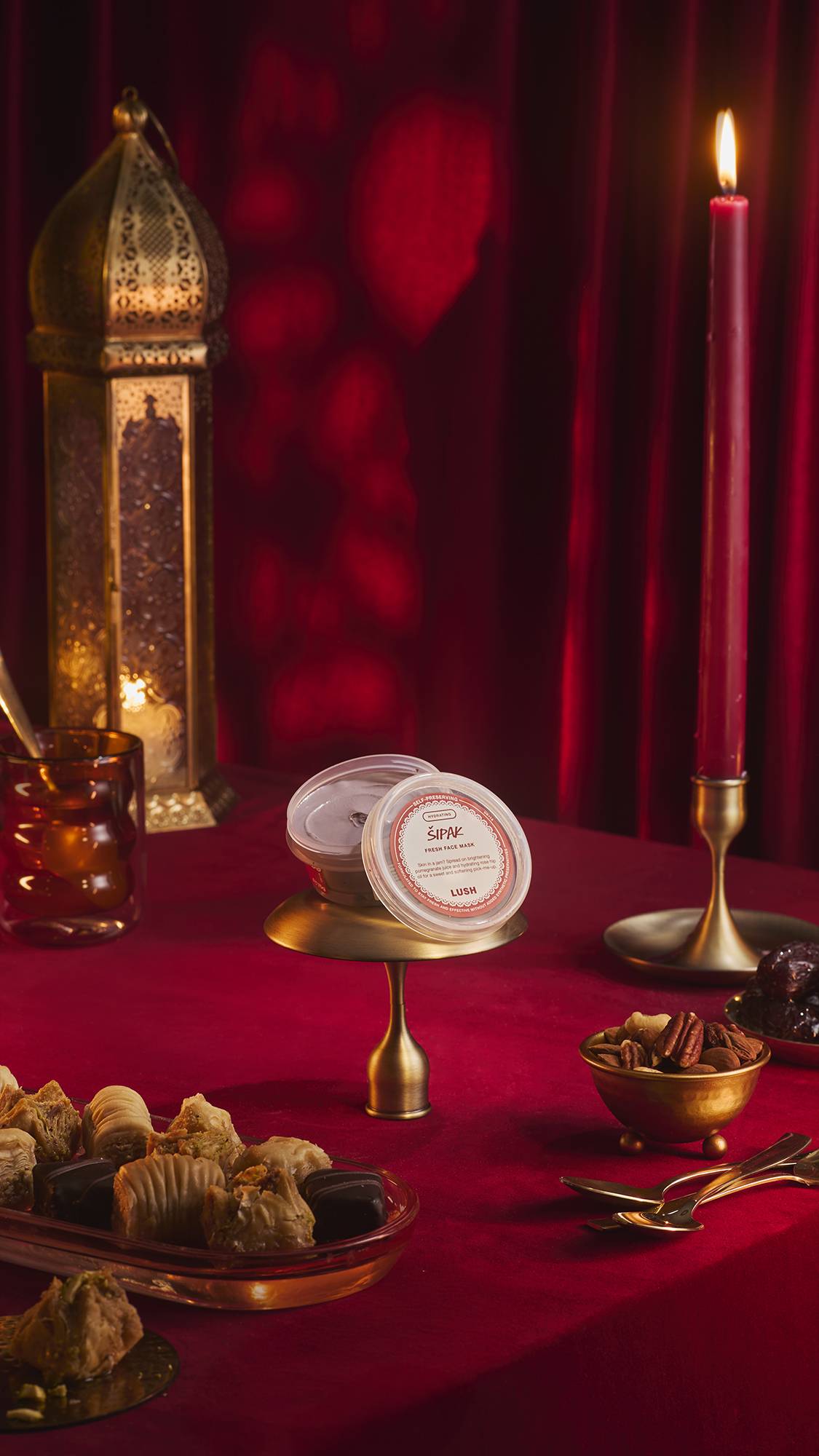 A pot of Lush 'Šipak' fresh face mask sits on a small brass stand atop a red velvet tablecloth. The festive scene features a tall gold lantern, a lit red candle, and bowls of nuts and pastries against a backdrop of draped red fabric.