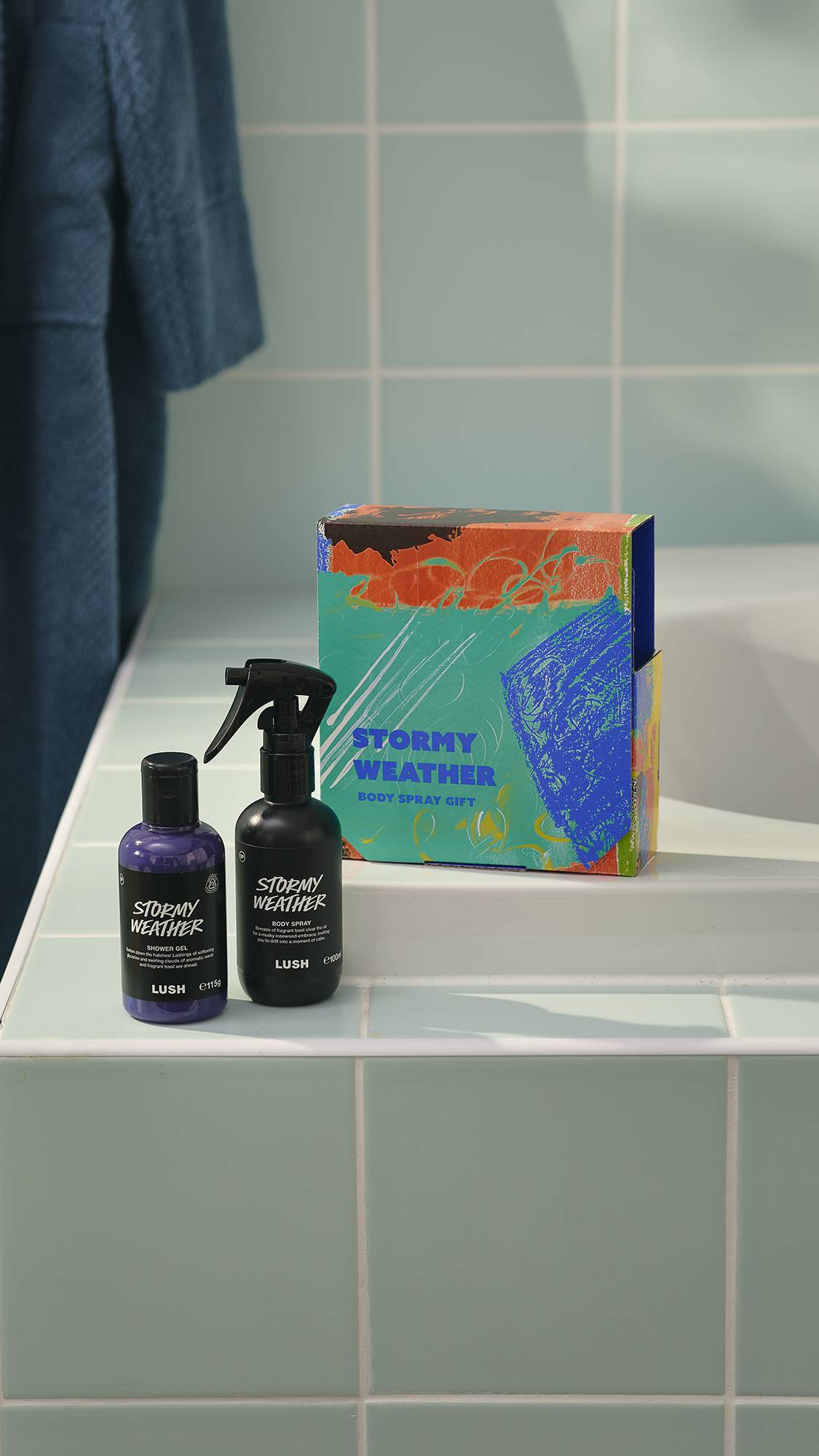 A Lush 'Stormy Weather' gift set sits on the edge of a bath against teal tiles. The set includes a colourful box, a bottle of purple shower gel, and a black bottle of body spray, both of the same name. A blue dressing gown hangs in the background.