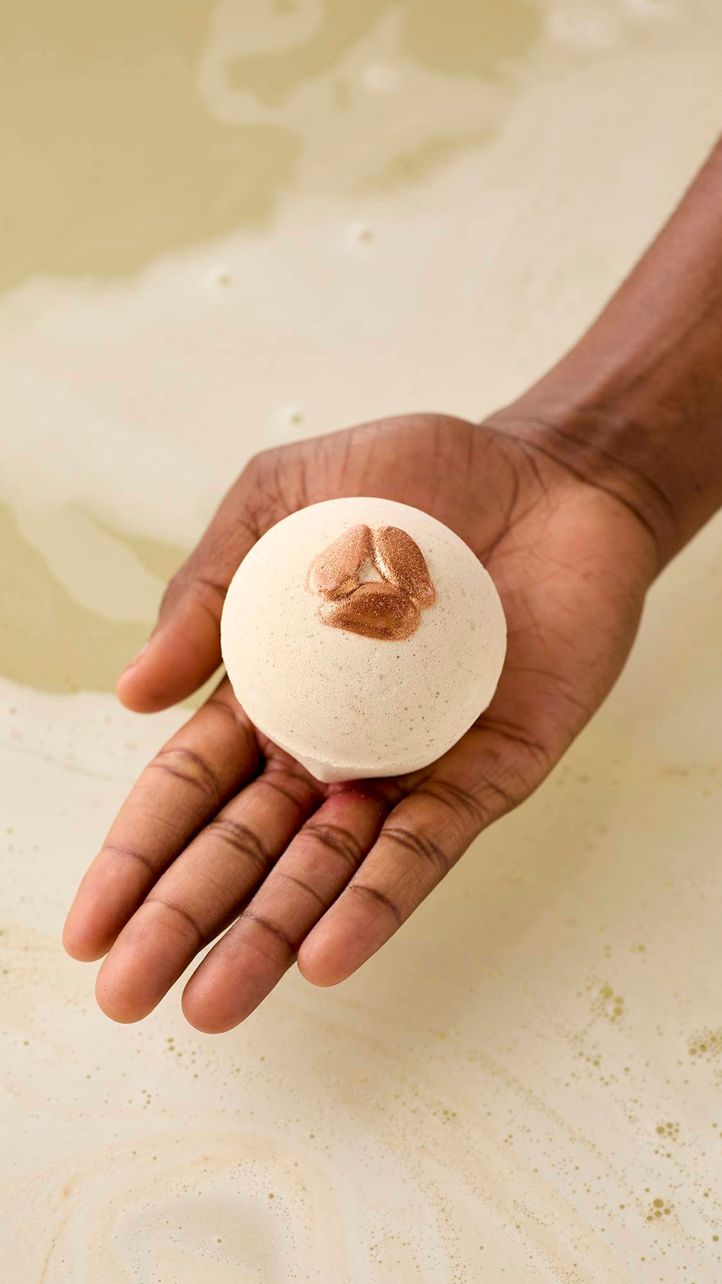 A hand holds a speckled, off-white Super Milk bath bomb with shimmering embossed almonds over a tub of pale, frothy water. The bath bomb is perfectly spherical, held in an open palm against a backdrop of creamy, swirling bathwater.