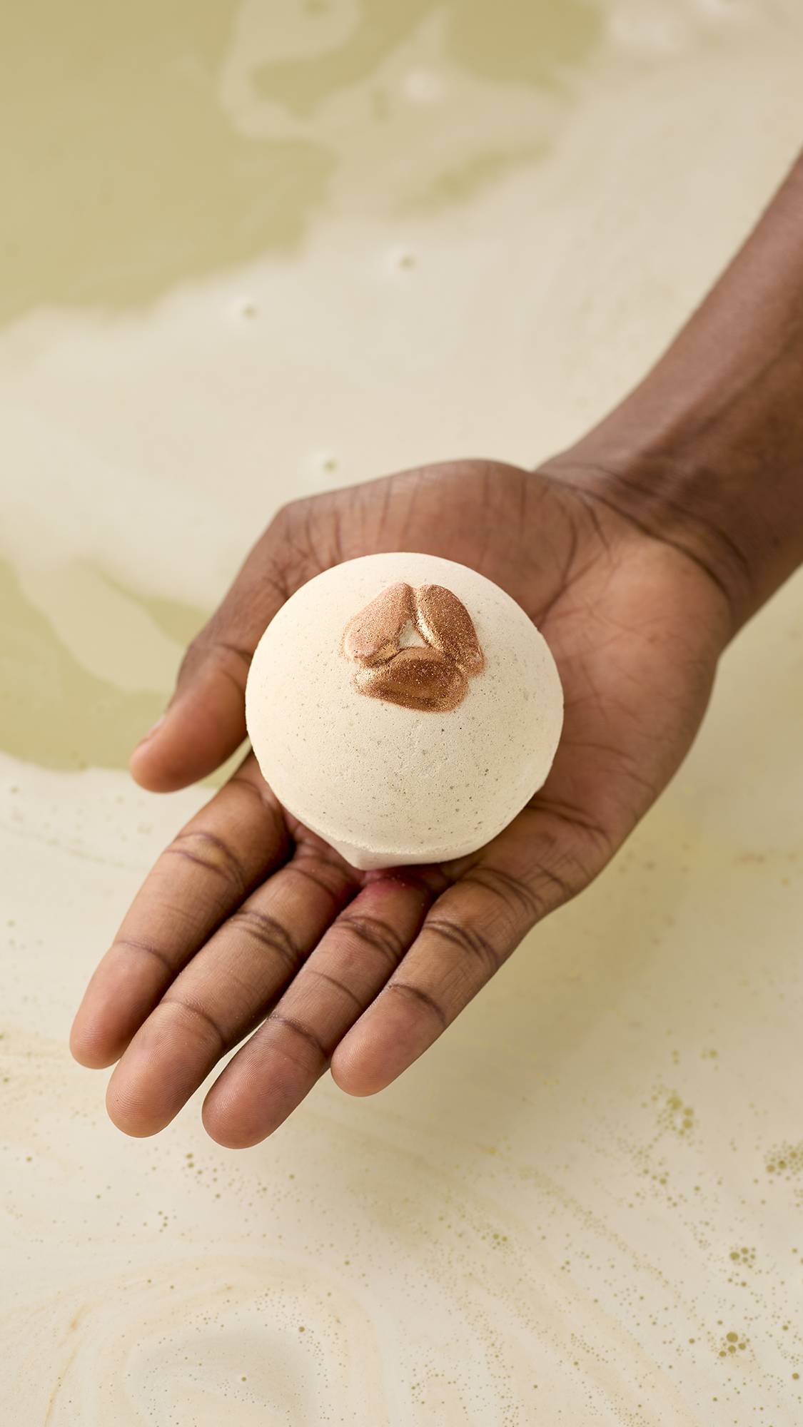 A hand holds a speckled, off-white Super Milk bath bomb with shimmering embossed almonds over a tub of pale, frothy water. The bath bomb is perfectly spherical, held in an open palm against a backdrop of creamy, swirling bathwater.