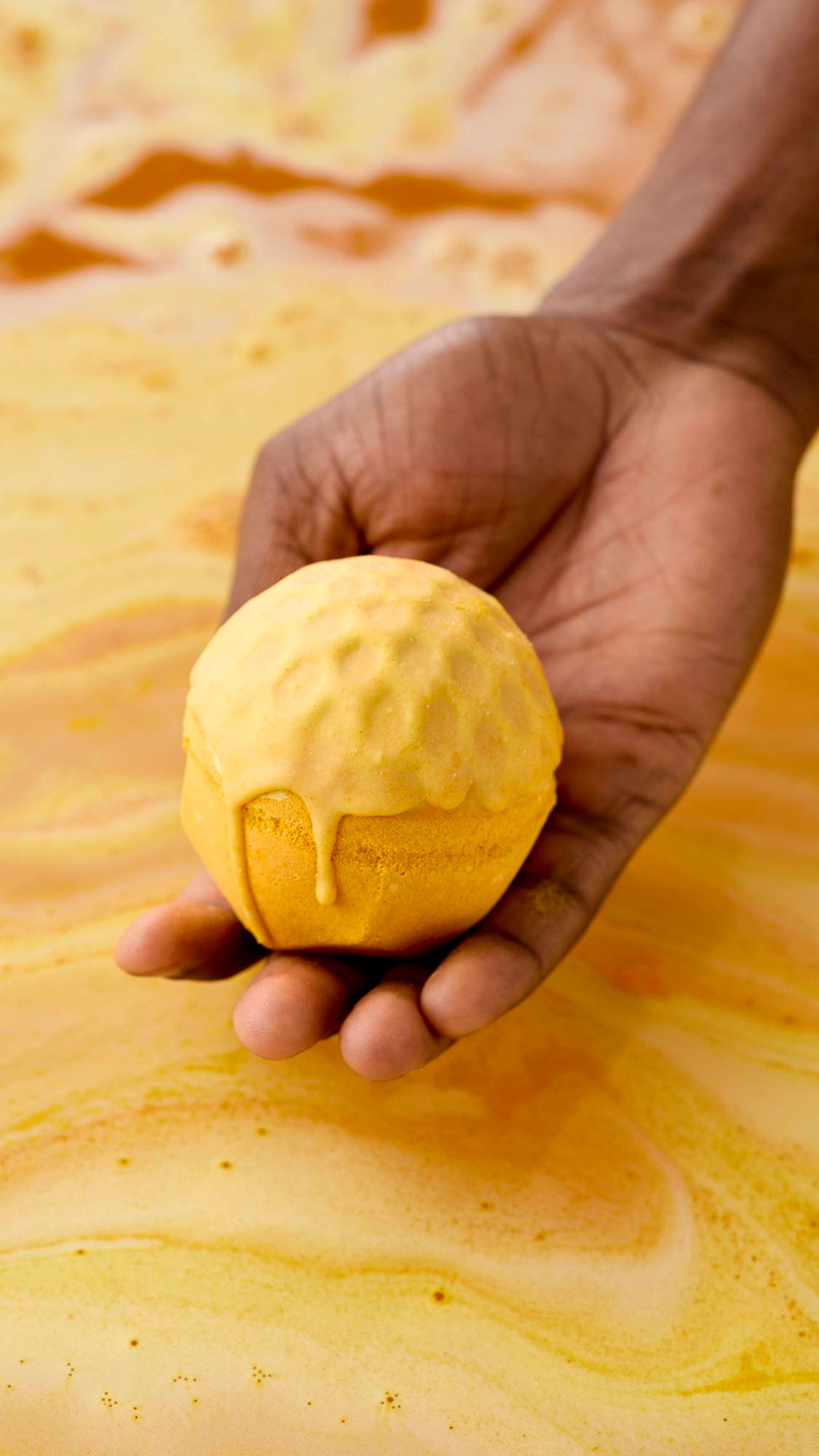 A hand holds a golden-yellow, "Sweet As Honey" bath bomb over a tub of swirling, amber-coloured water. The top of the bath bomb appears to be melting or dripping down the sides, set against the vibrant orange bathwater background.
