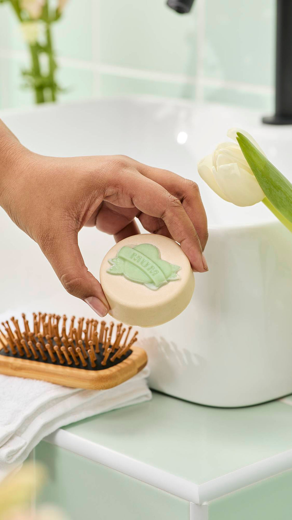 A hand holds a cream-coloured "Symbol of Love" solid oil bar with a green "MUM" banner design. It is displayed in a bathroom with a sink, wooden hairbrush, and white tulip against mint green tiles.