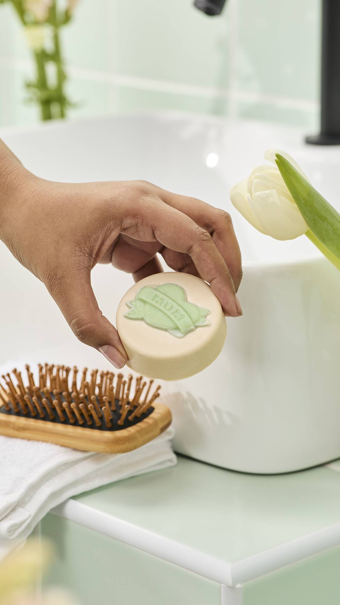 A hand holds a cream-coloured "Symbol of Love" solid oil bar with a green "MUM" banner design. It is displayed in a bathroom with a sink, wooden hairbrush, and white tulip against mint green tiles.