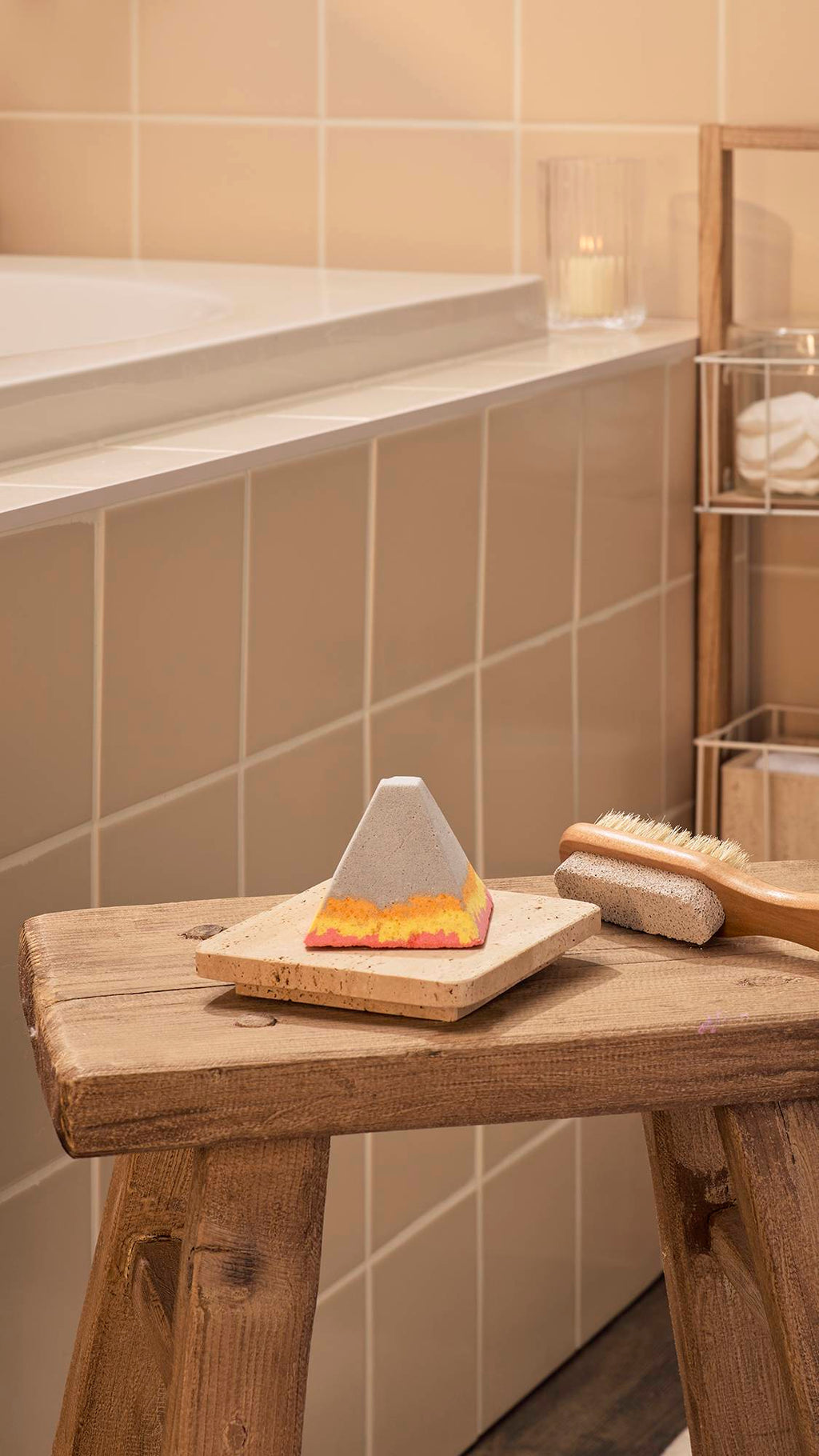 The "Volcano", a pyramid-shaped bath bomb-style foot soak with a grey top and wavy layers of orange, yellow, and pink at the base, sits on a stone tray positioned on a rustic wooden stool in a bathroom with neutral wall tiles and a lit candle.