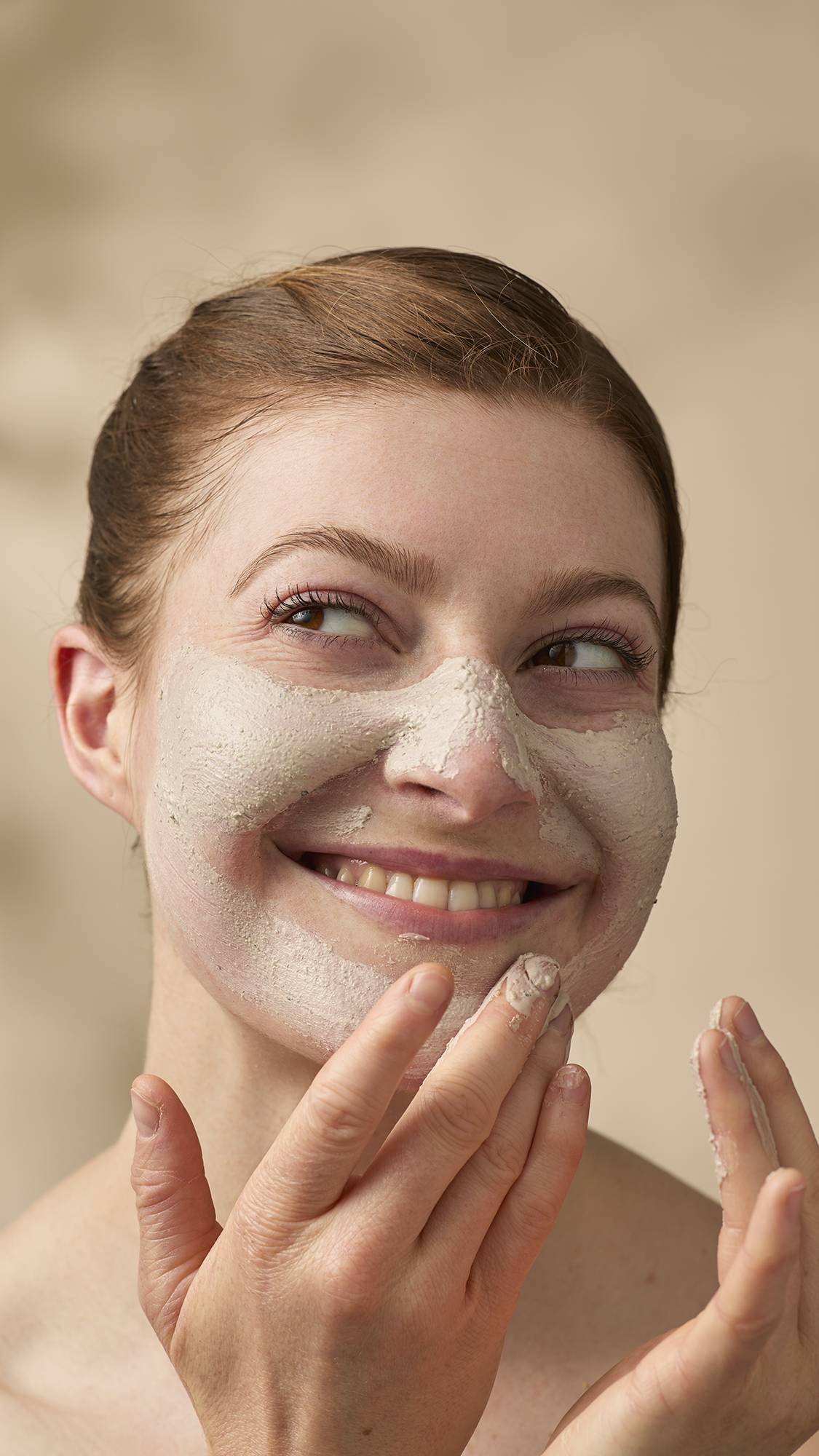 A close-up of a smiling model with reddish-brown hair tied back, applying a textured, beige face mask. They look off-camera with a playful expression while using their fingers to blend the thick cleanser across their nose, cheeks, and chin.