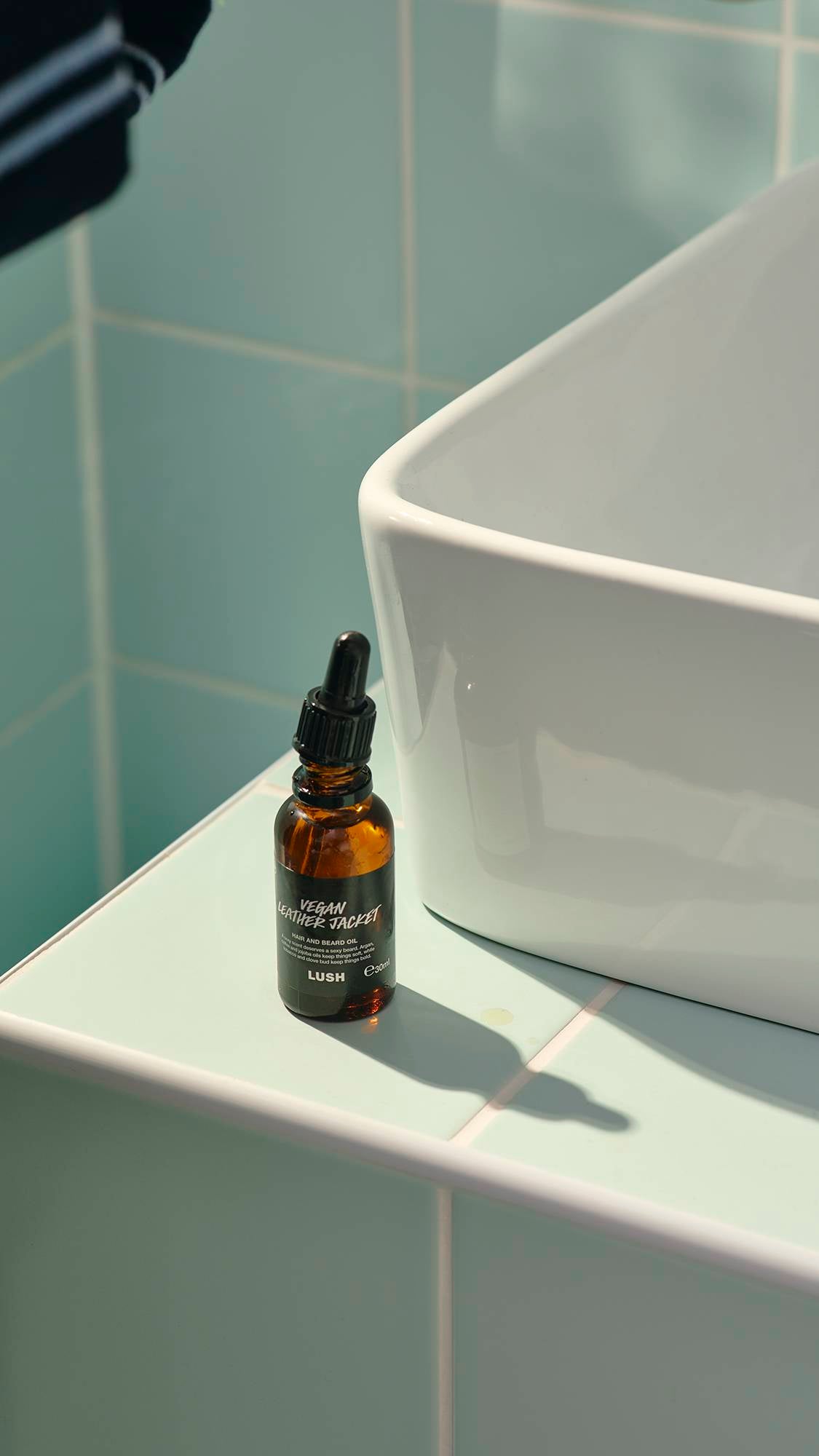 A small, amber glass bottle of "Vegan Leather Jacket" hair and beard oil by Lush sits on a pale green tiled ledge next to a white ceramic sink. The bottle features a black label and a pipette dropper lid, casting a soft shadow on the tiles.