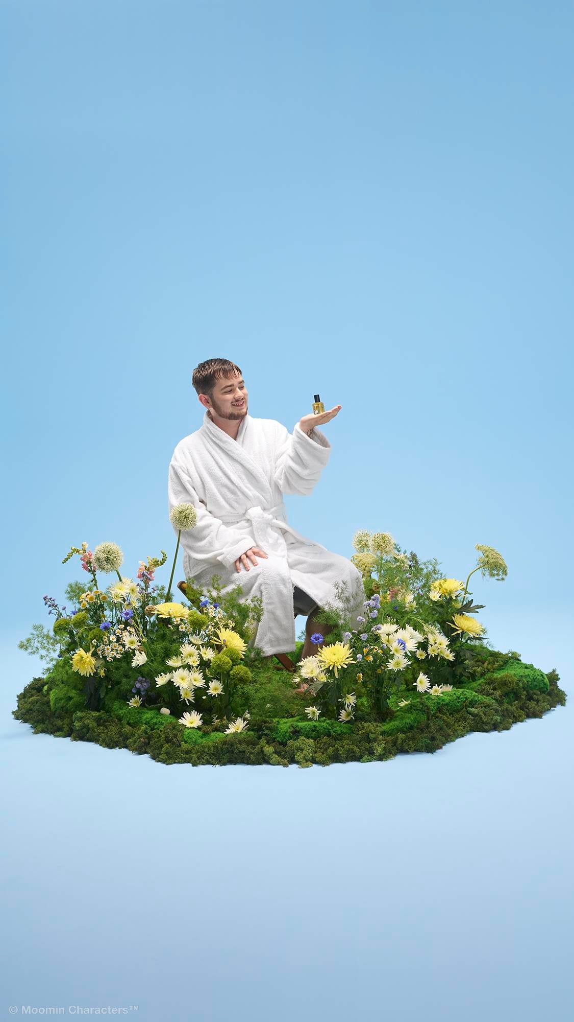 A person in a white dressing gown sits on a small patch of mossy ground surrounded by yellow and white wildflowers. They smile whilst holding a small "Moominvalley" perfume bottle in their open palm against a plain light blue background.