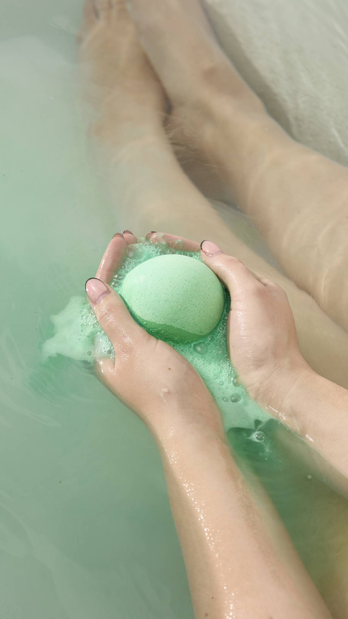 Hands hold the bright green Avobath Bath bomb as it fizzes in a tub of pale green water. A stream of frothy, light green bubbles flows from the bomb, with submerged legs visible in the background.