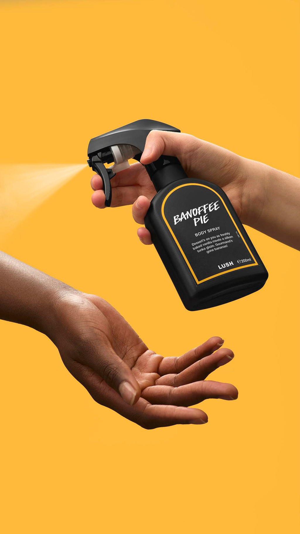 A person's hand presses the nozzle of a black Lush 'Banoffee Pie' body spray bottle, releasing a fine mist onto another person's open palm. The action is set against a solid, vibrant yellow background.