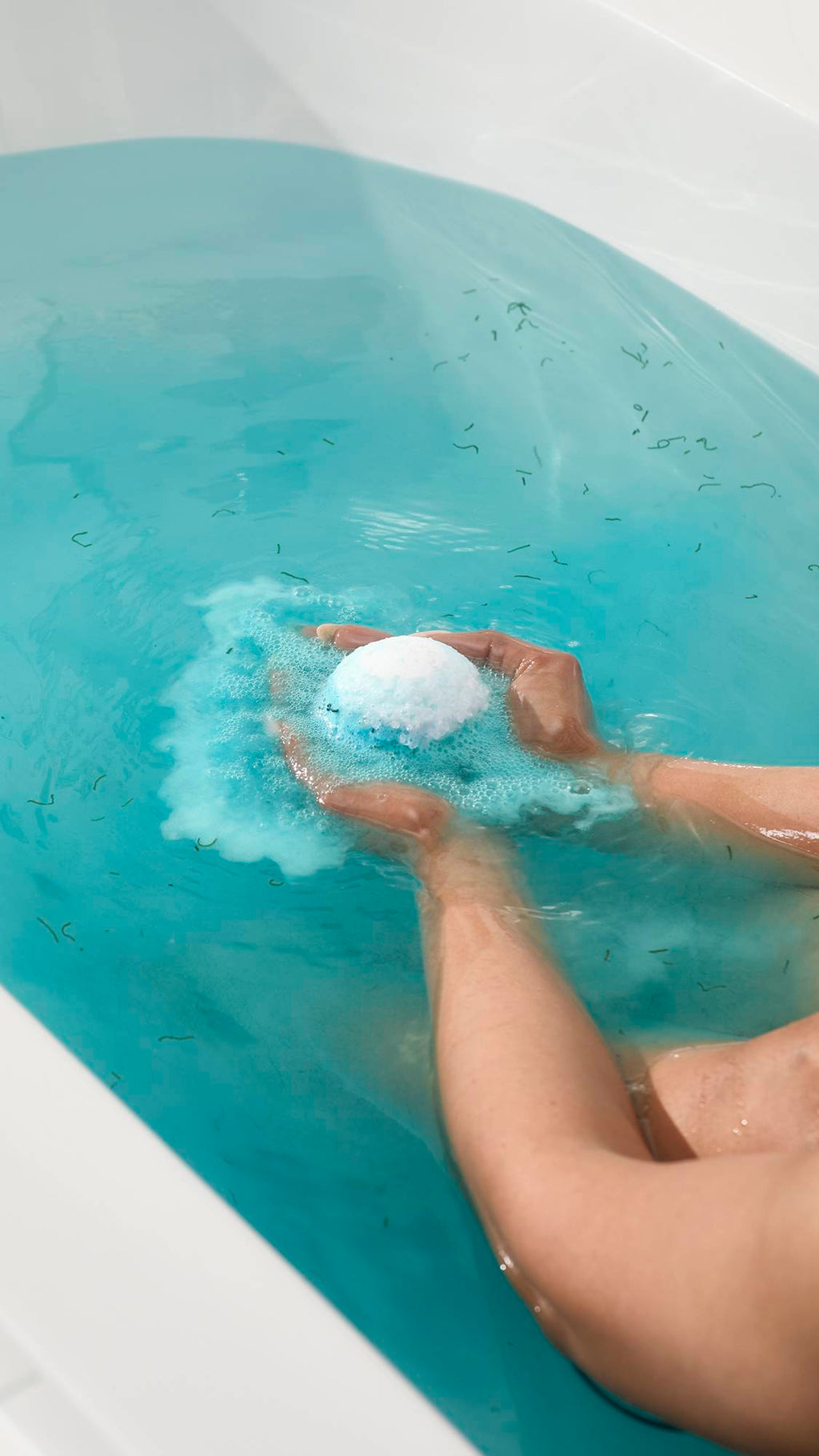 Hands hold the Big Blue bath bomb as it fizzes in a tub of bright blue water. A cloud of frothy blue foam surrounds the bomb, while small green pieces of seaweed float throughout the water.