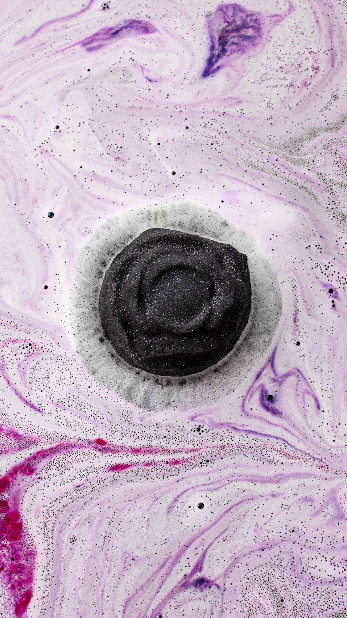 Black Rose. An aerial view shows the dark rose-shaped bath bomb as it dissolves. It creates a thick layer of velvety, deep-purple foam on the water's surface, rippling with bright pink swirls and shimmering silver plastic-free glitter.