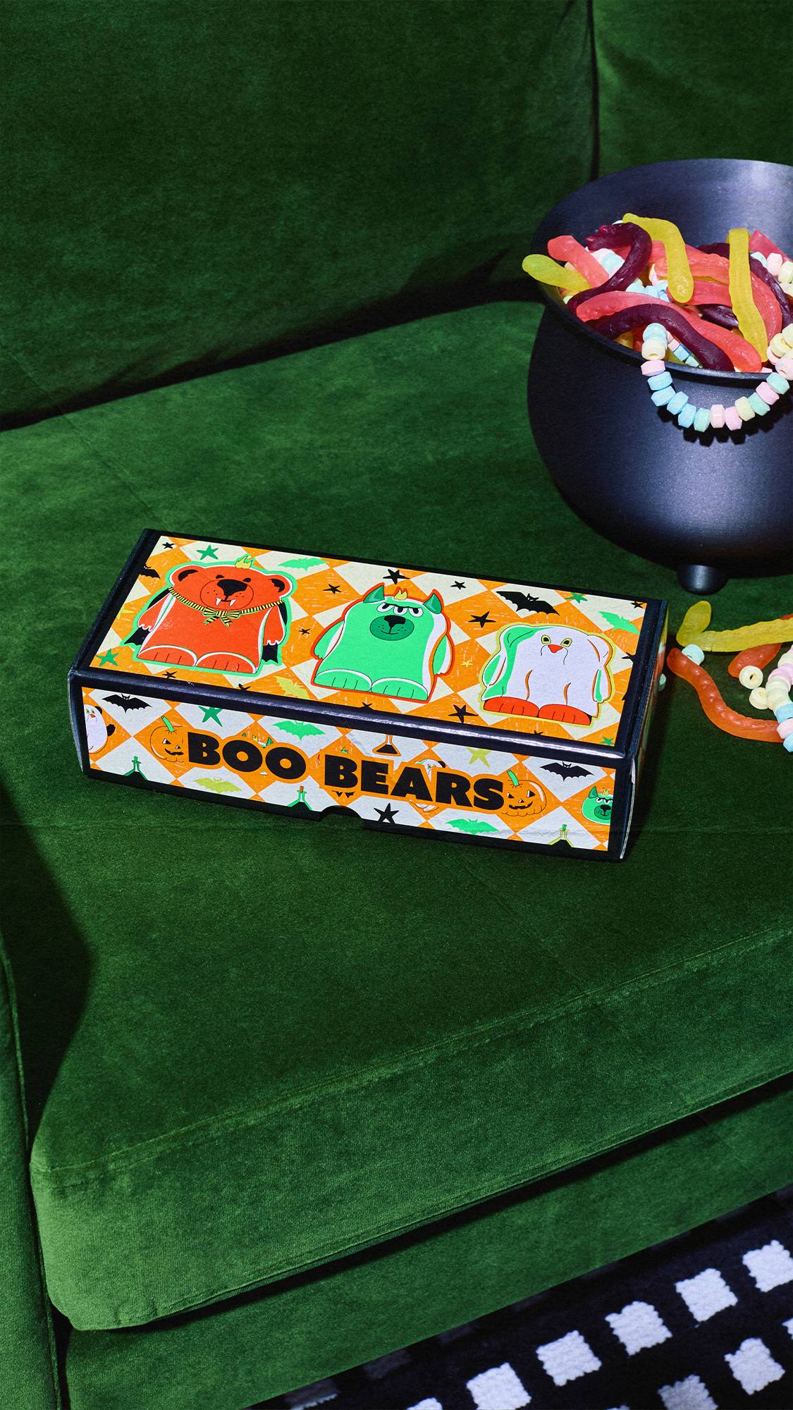 A Lush 'Boo Bears' gift box with orange chequered patterns and monster illustrations sits on a green velvet couch. Next to it, a black cauldron is filled with colourful sweets like gummy worms and candy necklaces.