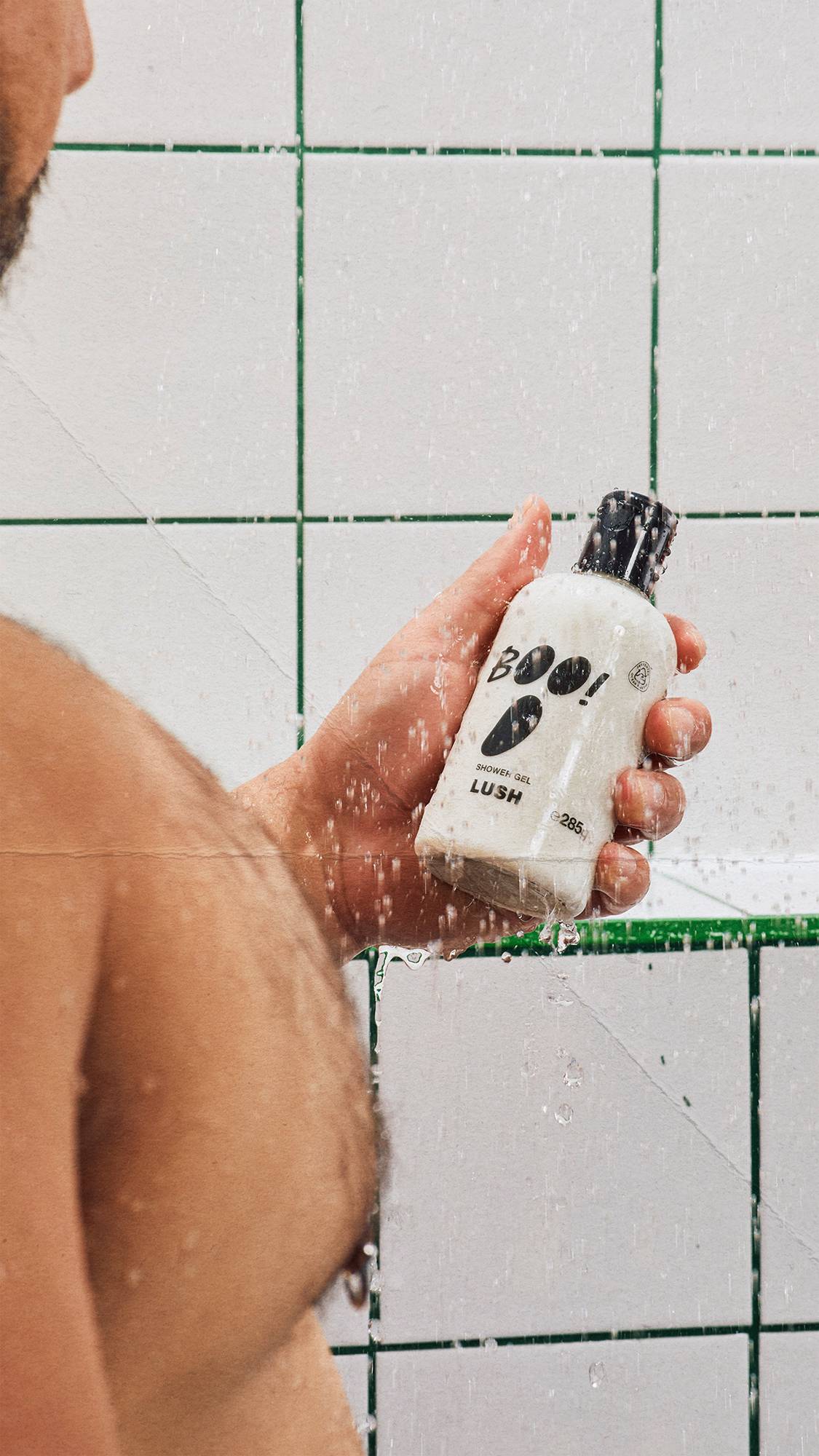 A model holds a bottle of Lush 'BOO!' shower gel under streaming water in the shower. Water droplets splash over the bottle, which features a black ghost face design. White wall tiles with green grout form the background.