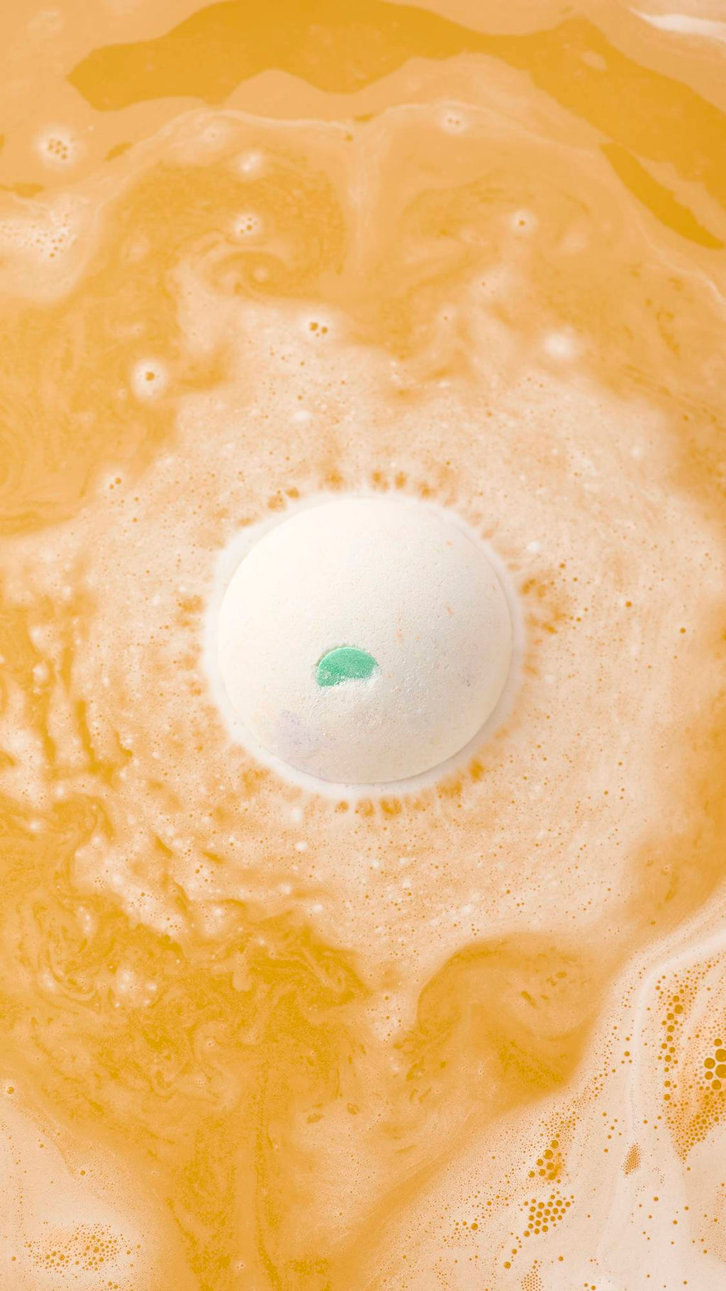 An aerial view of the "Dragon's Egg" bath bomb with a small green accent dissolving in vibrant orange water. It is surrounded by a ring of frothy white bubbles and swirling, marbled patterns across the surface.