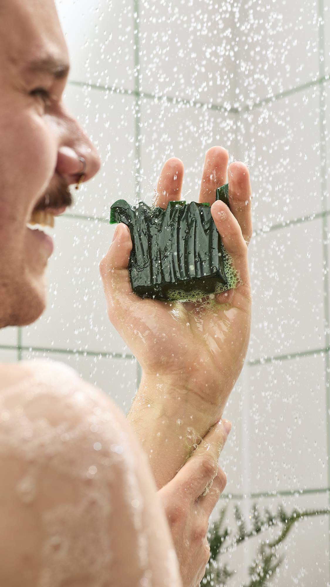 A smiling person in the shower holds a textured, dark green shower gummy soap in their hand. Water sprays around them against a backdrop of white tiles with green grout.