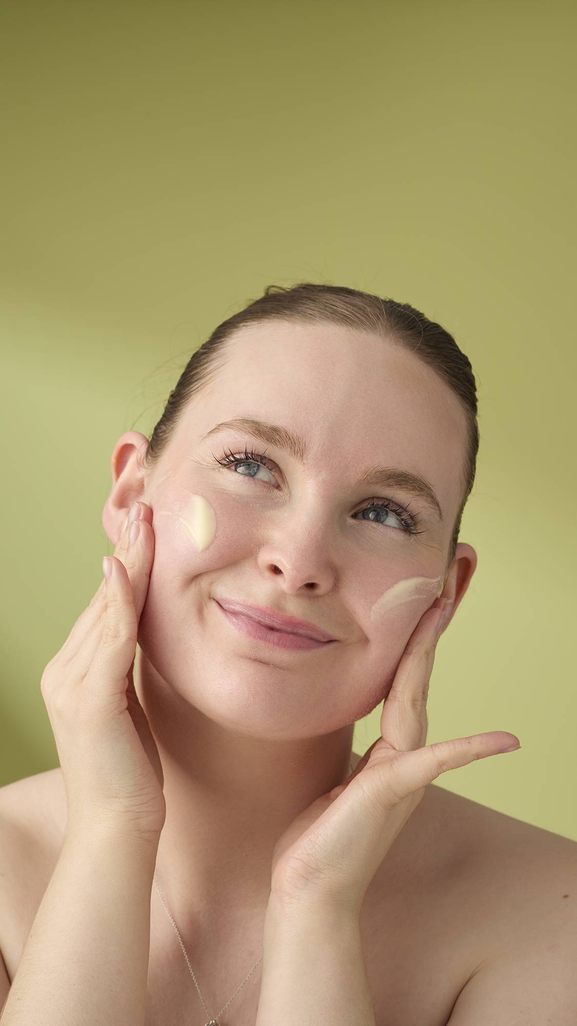 A model with light hair smiles while applying the yellow Honey, Beeswax And Almond Oil cleanser to her face. She is shown from the shoulders up against a solid olive-green background, highlighting the creamy texture of the facial product.