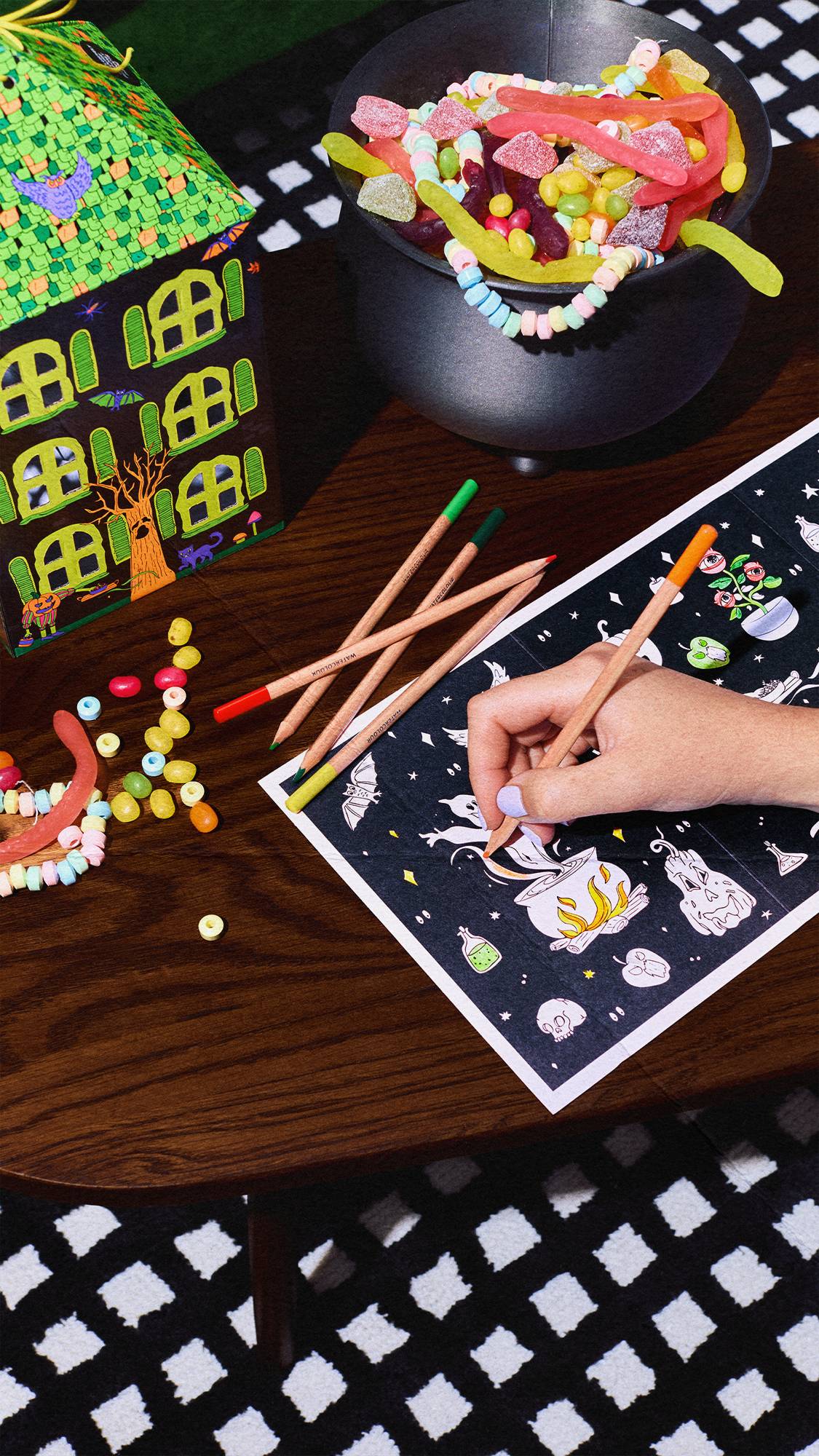 A hand uses a pencil to colour in a Halloween sheet on a wooden table. A house-shaped gift box, loose coloured pencils, and a black cauldron filled with colourful sweets sit nearby.