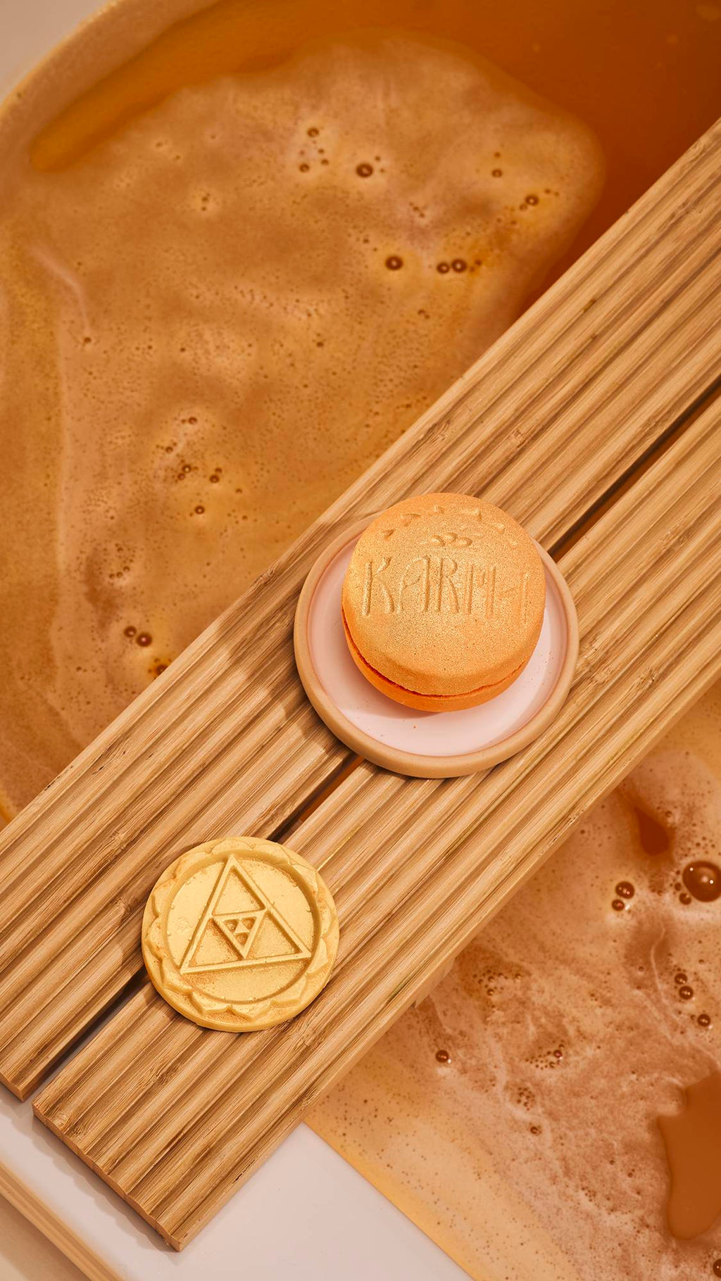 An orange, macaron-shaped 'Karma' bath bomb rests on a ceramic dish. Beside it on a wooden bath tray is the pale-yellow, circular middle layer embossed with a triangle design. Below, the bathwater is a deep amber colour with a thick layer of foam.