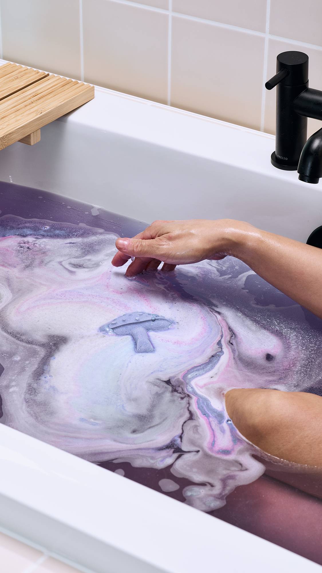 The image shows the purple "Magik Mushroom" floating in a bath of deep lilac water. It is surrounded by swirls of white, pink, and pale blue foam as it dissolves. A model's arm reaches towards the floating mushroom from the side.