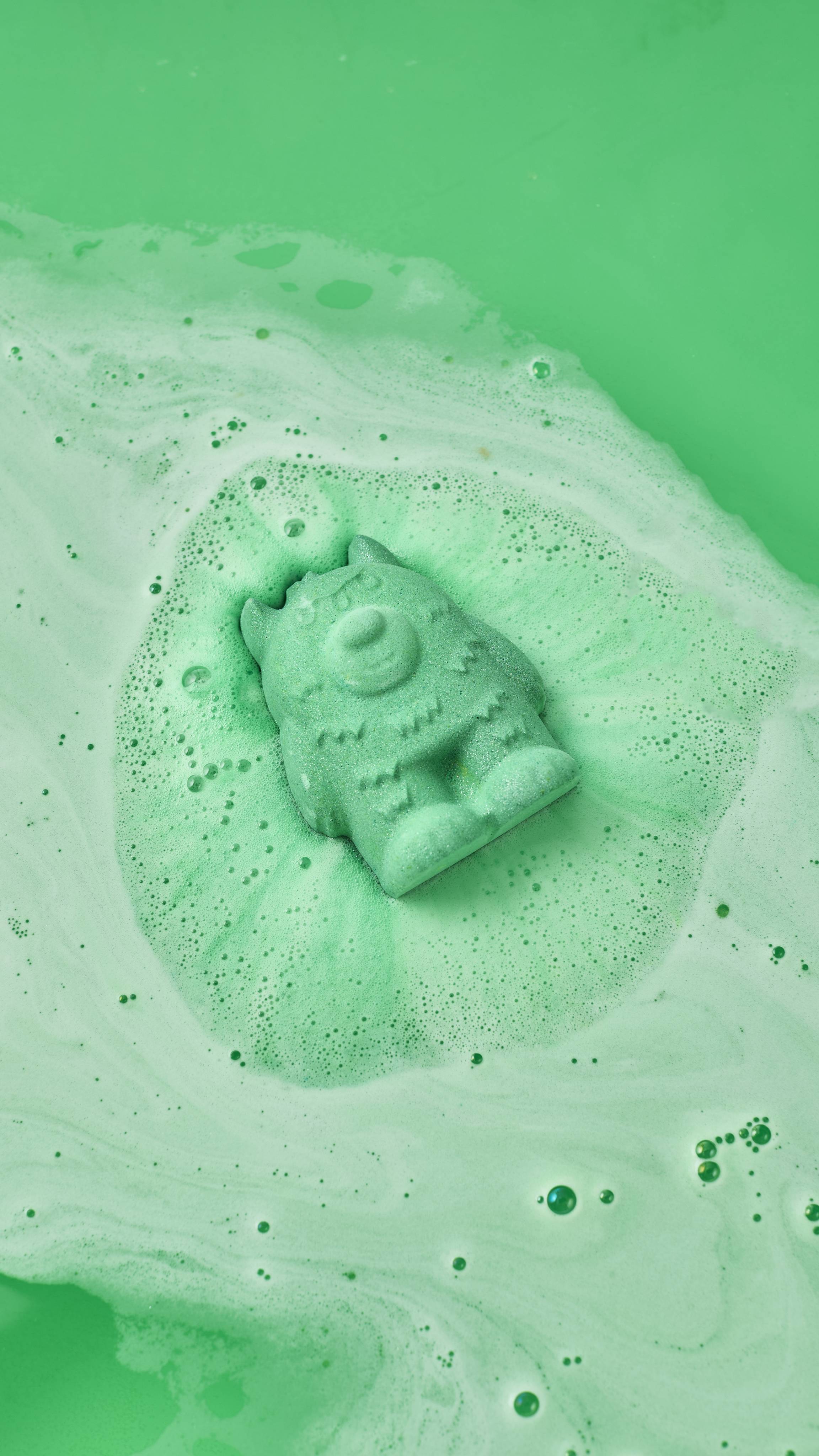 A green, three-eyed monster bath bomb fizzes and dissolves in bright green bathwater, surrounded by swirls of light green foam and bubbles.