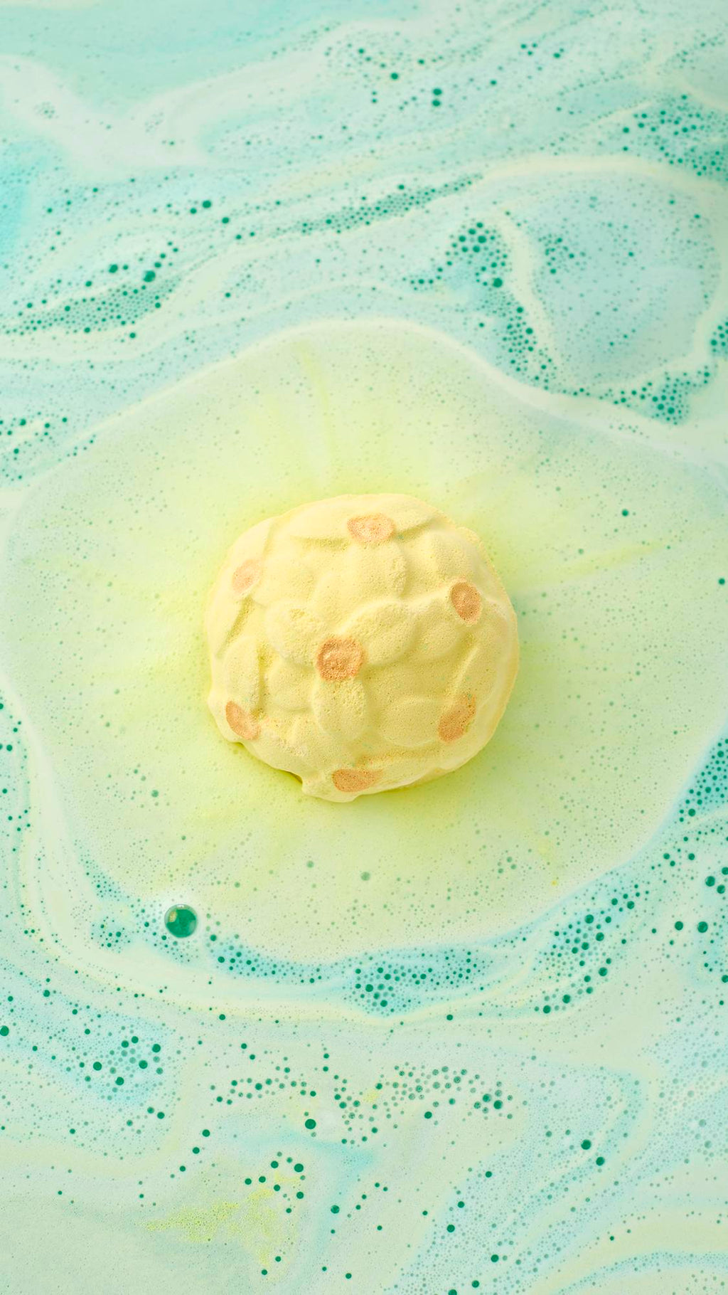 A top-down view of the yellow Narcissus bath bomb floating in shimmering green water. It fizzes with a bright yellow center, surrounded by soft swirls of pale green and white foam that create a marbled effect across the bath.