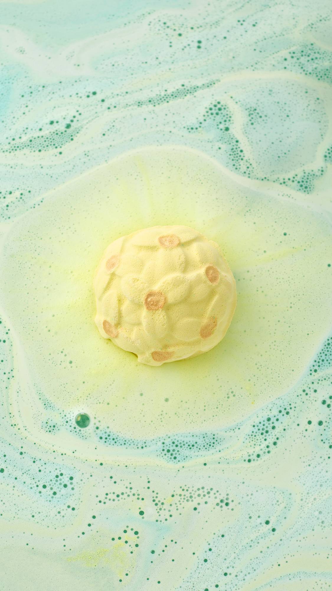 A top-down view of the yellow Narcissus bath bomb floating in shimmering green water. It fizzes with a bright yellow center, surrounded by soft swirls of pale green and white foam that create a marbled effect across the bath.