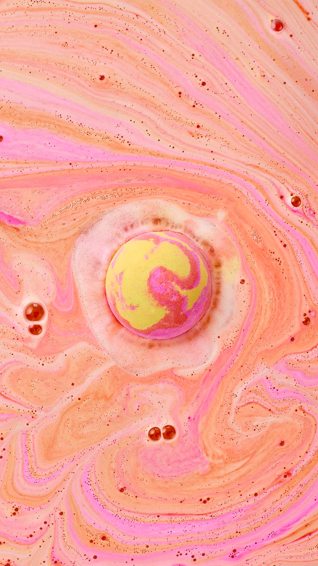 A top-down view of the Passion bath bomb floating in a bath, swirling with vibrant pink and yellow foam. The water is a warm peach color, filled with intricate, marble-like patterns of neon pink and soft orange bubbles.