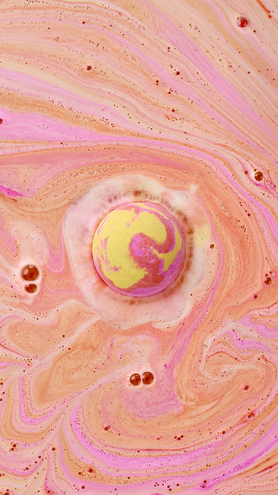 A top-down view of the Passion bath bomb floating in a bath, swirling with vibrant pink and yellow foam. The water is a warm peach color, filled with intricate, marble-like patterns of neon pink and soft orange bubbles.