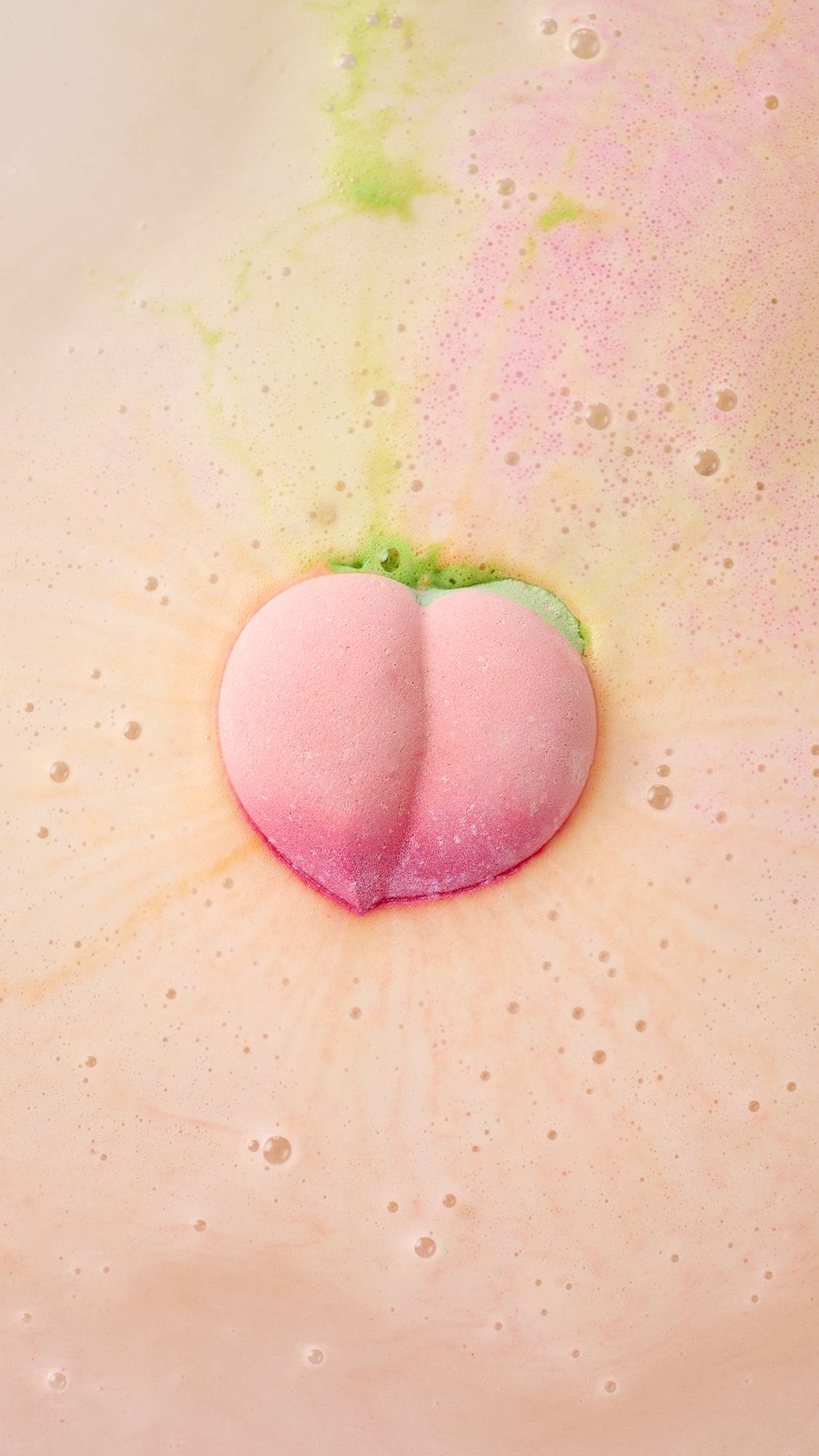 A peach-shaped bath bomb fizzes in the centre of a tub, releasing swirls of vibrant pink and lime green into creamy, peach-coloured water. The surface is covered in a delicate layer of bubbles and frothy foam.