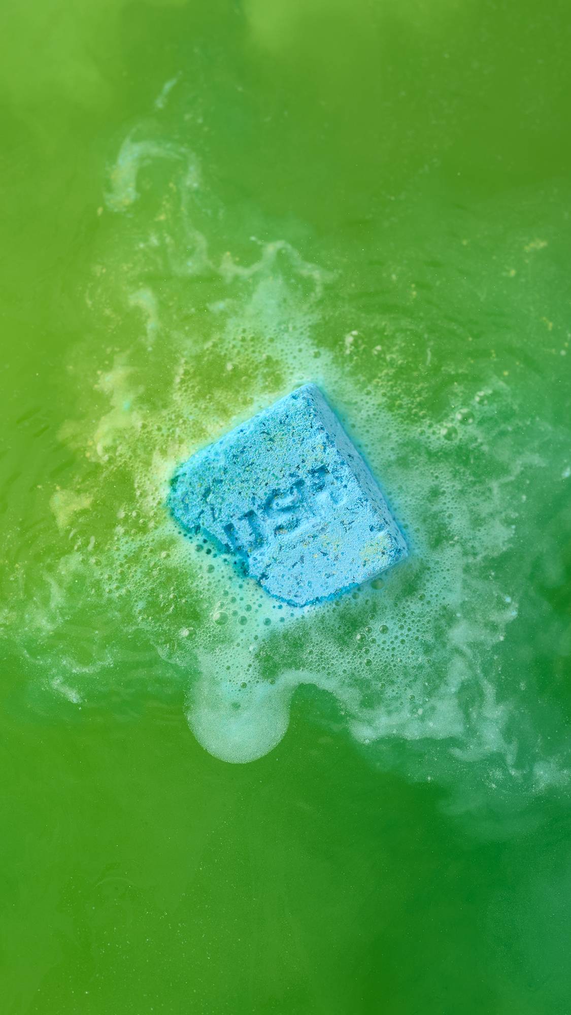 The blue and yellow, cubed "Salt Water Soother" bath bomb, with "LUSH" debossed on its surface, dissolves in bright green bathwater. It is surrounded by a patch of white, frothy bubbles as the vibrant blue and green colours begin to swirl together.