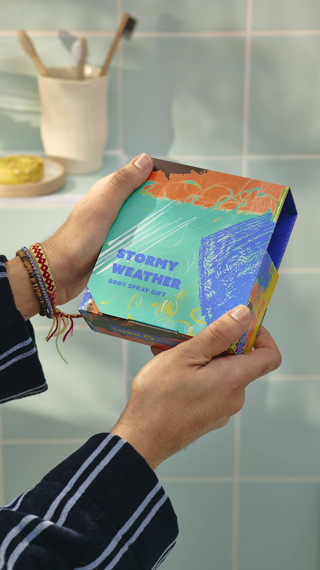 A model wearing a striped dressing gown holds a Lush ‘Stormy Weather’ gift box. The square box features a vibrant, abstract design in teal, orange, and blue. In the background, toothbrushes sit in a cup against blue bathroom tiles.