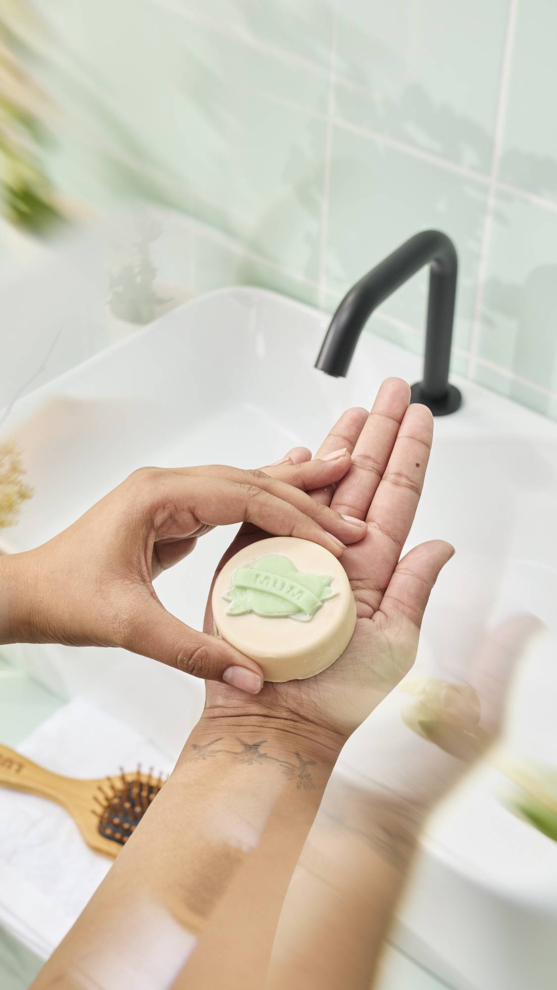 Hands hold a round, cream-coloured "Symbol of Love" body oil bar featuring a green heart and banner design reading "MUM" over a bathroom sink. A tattoo of three flying birds is visible on the wrist against a mint green tiled background.