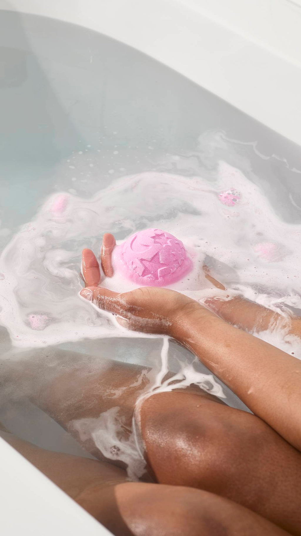 Lavender Twilight Bath Bomb with star and moon designs fizzes in hands over white bathwater foam.