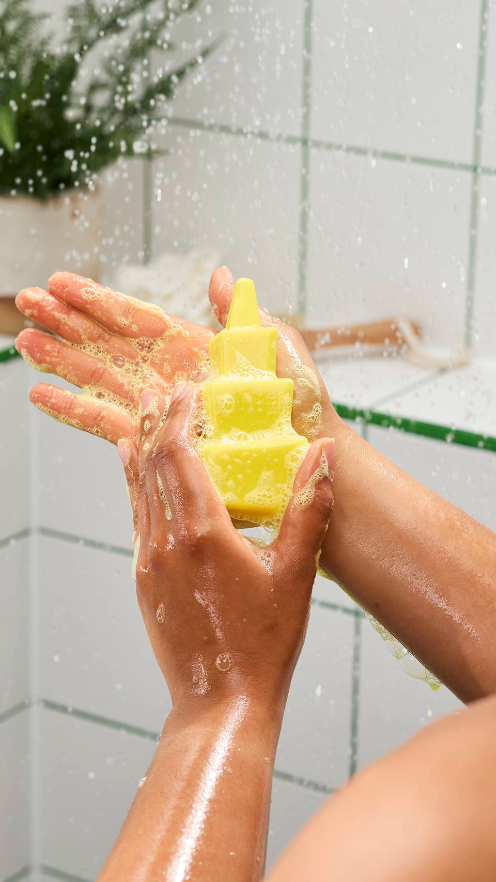 Hands lathering a bright yellow, tiered geometric shower gummy soap under running water. Bubbles cover the hands against a background of white tiles with green grout and splashing water.