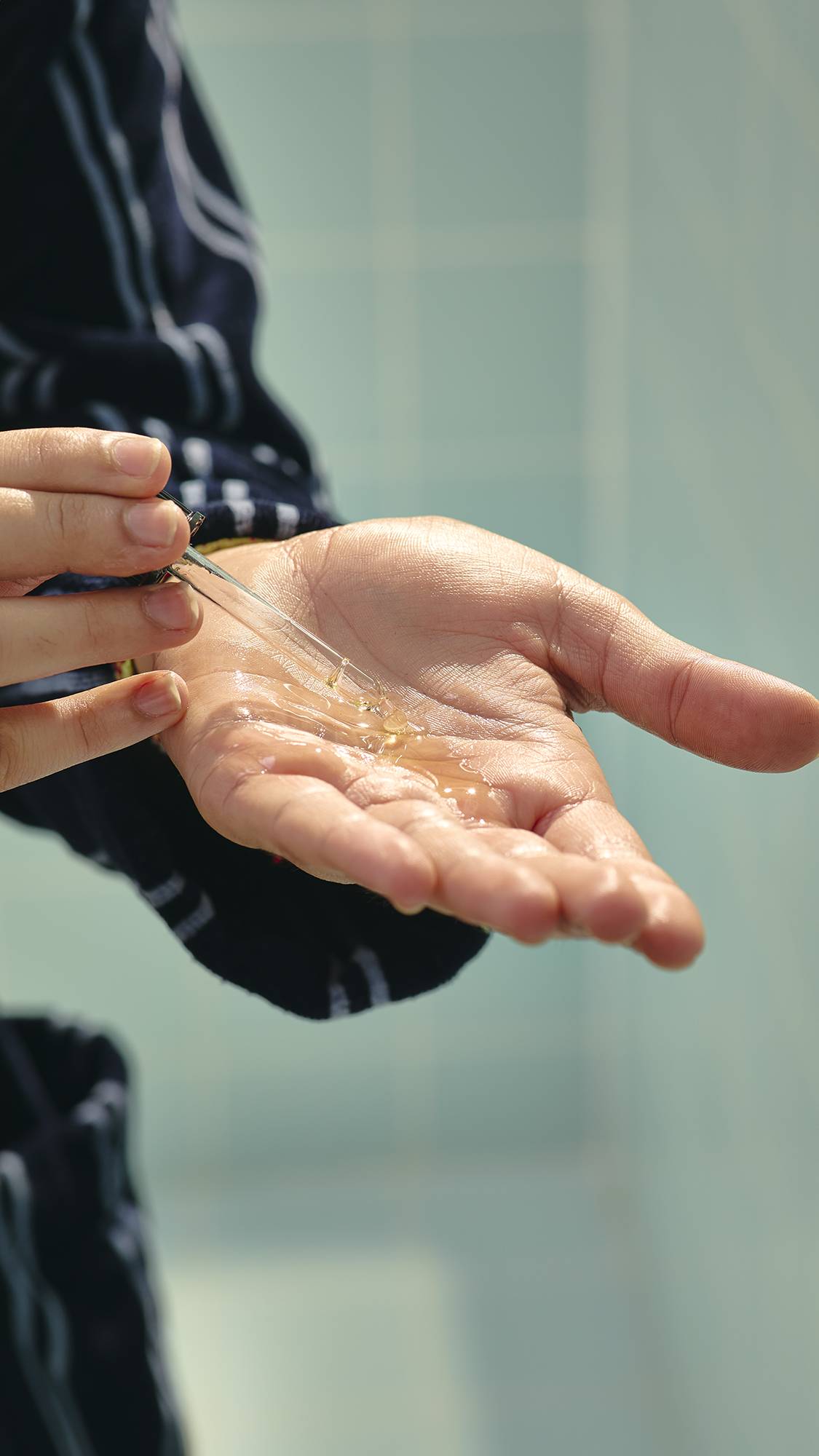 The image shows a close-up of a model's hands as they hold the glass bottle of "Vegan Leather Jacket" hair and beard oil and use the pipette topper to drop clear, golden oil into their open palm.