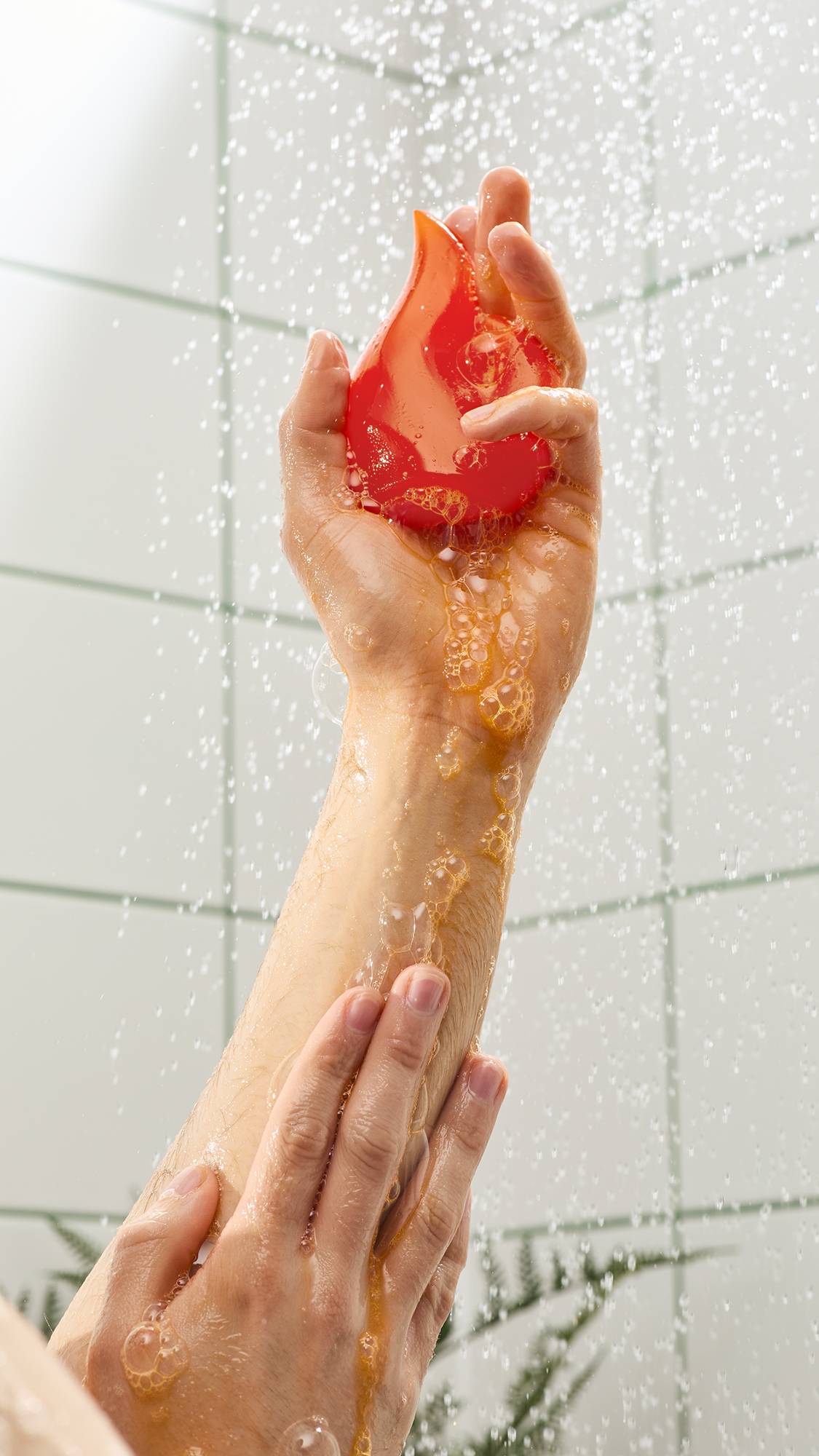 A hand holds a bright orange, flame-shaped shower gummy soap under spraying water. An orange lather with bubbles runs down a wet forearm against a background of white tiles with green grout.